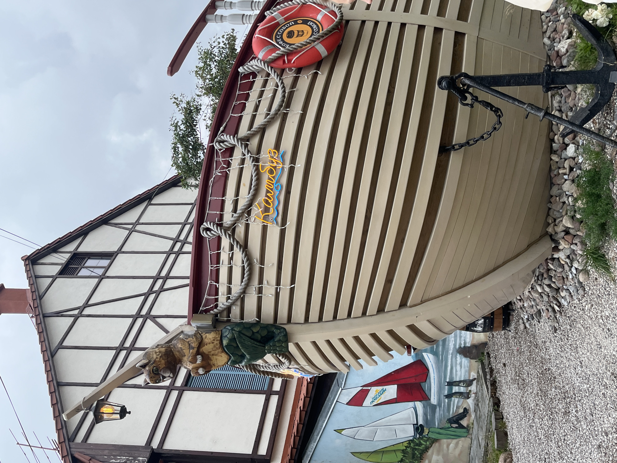 This image depicts the exterior of a building with a boat-themed facade in Zelenogradsk, Russia. The building has a half-timbered architectural style with a red tiled roof and a chimney. Attached to the front of the building is a large, beige, boat-shaped structure with horizontal planks. A wooden carving of a cat with a mermaid tail is mounted on the bow of the boat structure, next to an old-fashioned lantern. There are ropes and string lights decorating the top edge of the boat. A red life preserver ring hangs on the side of the boat. A neon sign with Cyrillic text reading "Камбуз" (meaning "galley") is visible. Below the boat structure, a mural of sailboats on the sea is painted on a wall. The ground in front of the building is covered with gravel and stones. An anchor is placed in front of the boat structure, secured by a chain. The sky is overcast.