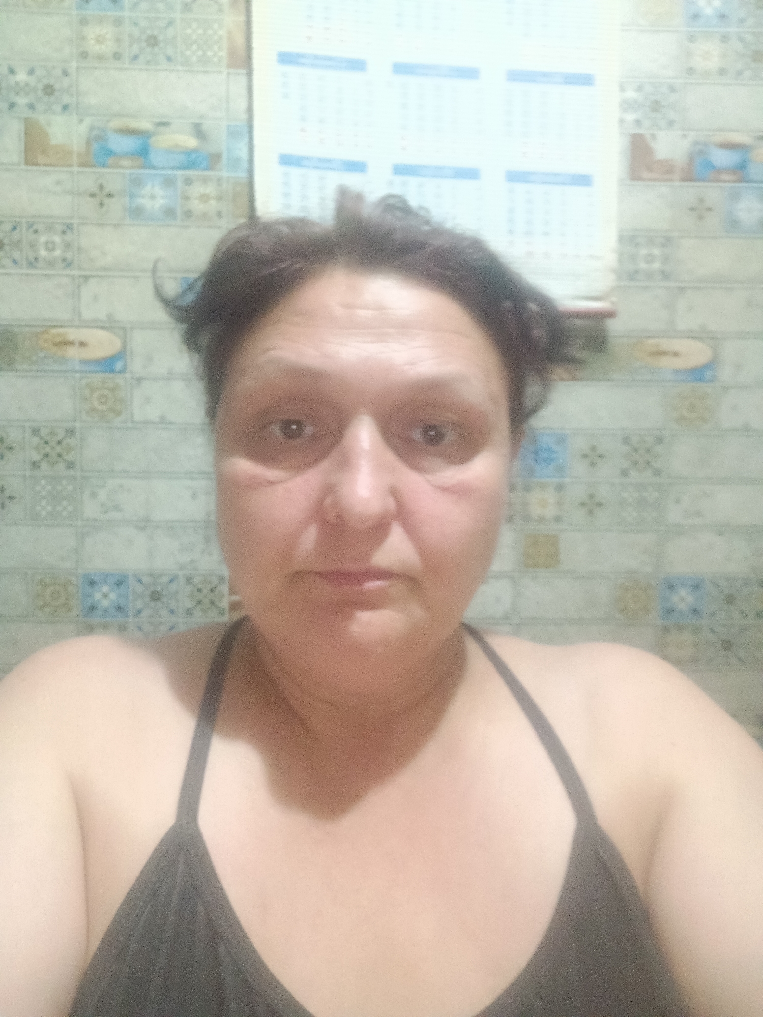 This is a portrait of a woman in Kokshetau, Kazakhstan. The woman has short, dark hair and appears to be in her late 40s or early 50s. She is wearing a dark-colored tank top. The background consists of a tiled wall, with a calendar hanging on it. The lighting suggests an indoor setting, possibly a bathroom or kitchen. The image is slightly blurry, indicating it may have been taken with a mobile phone camera.