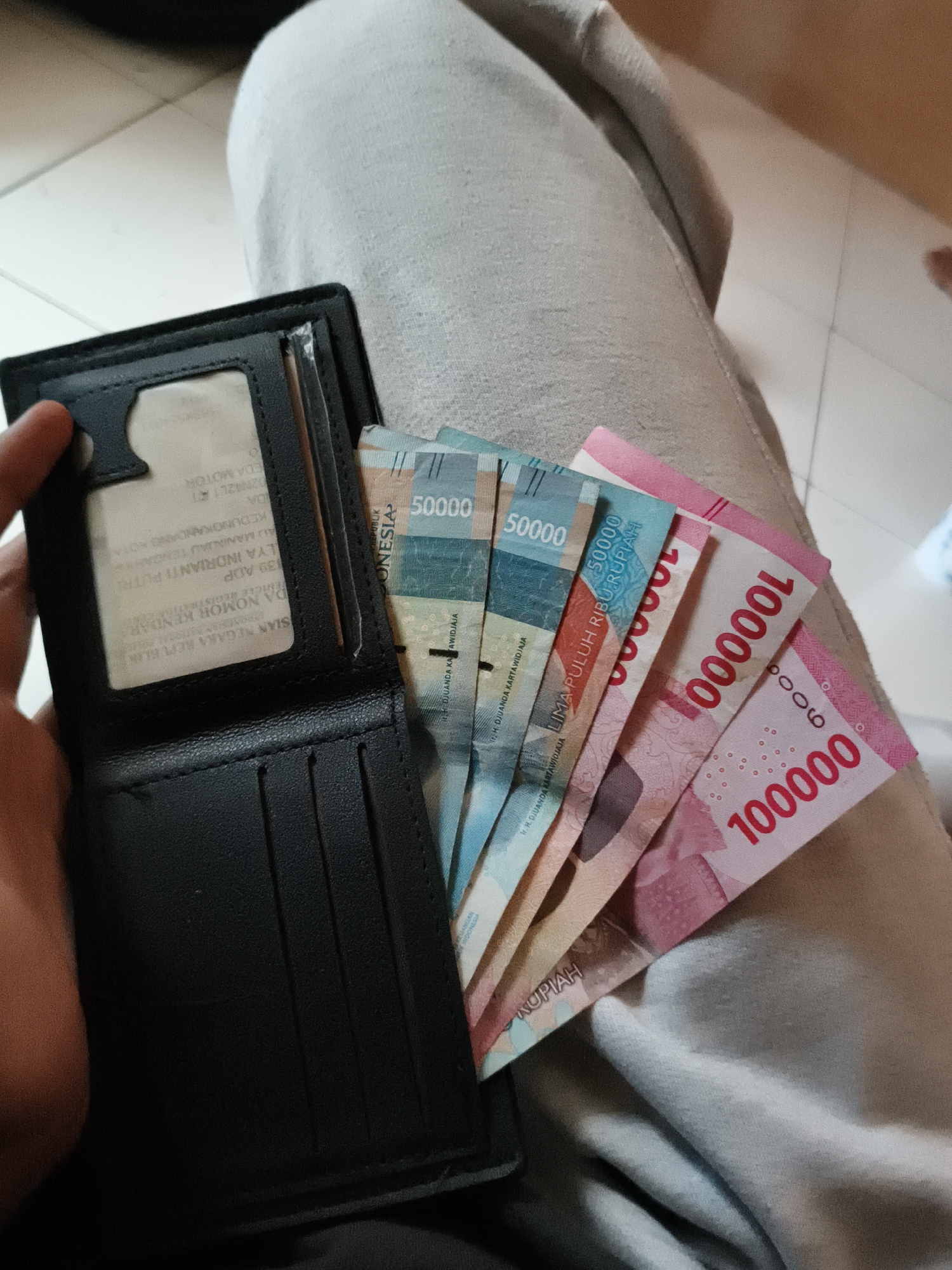 The image shows a person's hand holding a black wallet that is partially open. Inside the wallet, several Indonesian Rupiah banknotes are fanned out. The banknotes include denominations of 50,000 and 100,000 Rupiah. The wallet also contains what appears to be a vehicle registration document. The person is wearing light-colored pants. The background is blurred but suggests an indoor setting with tiled flooring. There are no discernible landmarks to indicate the specific location within Cianjur, Indonesia.