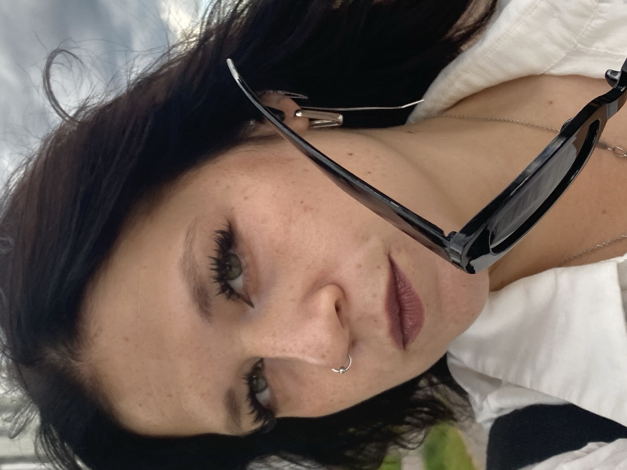 A woman with dark hair and green eyes is pictured in this medium shot. She is wearing a white collared shirt with a black strap visible over her shoulder. She has a nose ring and two earrings, one a silver square stud and the other a silver chain earring. She is holding a pair of black sunglasses in front of her face. The background appears to be an outdoor setting with a cloudy sky and some greenery visible on the left. The image does not provide any specific information to indicate the location within the city of Saint Petersburg, Russia.