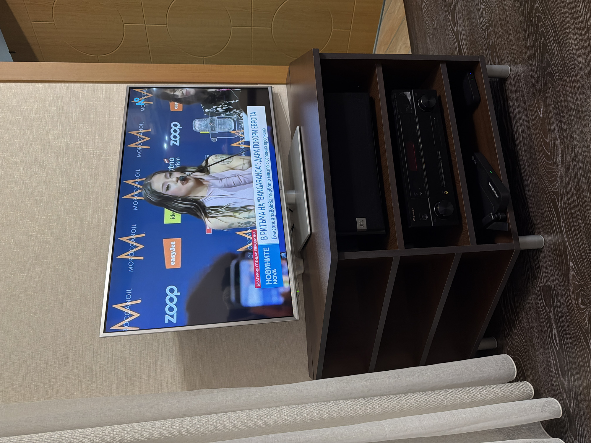 The image shows a television displaying a news broadcast. The broadcast is from NOVA, a Bulgarian television channel, and features information about Bulgaria winning the Eurovision Song Contest. The text on the screen includes "В РИТЪМА НА 'BANGARANGA': ДАРА ПОКОРИ ЕВРОПА" which translates to "In the rhythm of 'BANGARANGA': Dara conquers Europe." The screen also displays advertisements, including logos for Zoop, EasyJet, and Moroccanoil.
The television sits atop a dark brown shelf unit. On the shelves are a JBL soundbar, a Pioneer stereo receiver, and a VIVACOM router. The shelf unit is positioned on a wooden floor.
The lower portion of the frame includes a white, horizontally rolled-up blind or curtain. There is no information on the location, city or information about Burgas, Bulgaria.