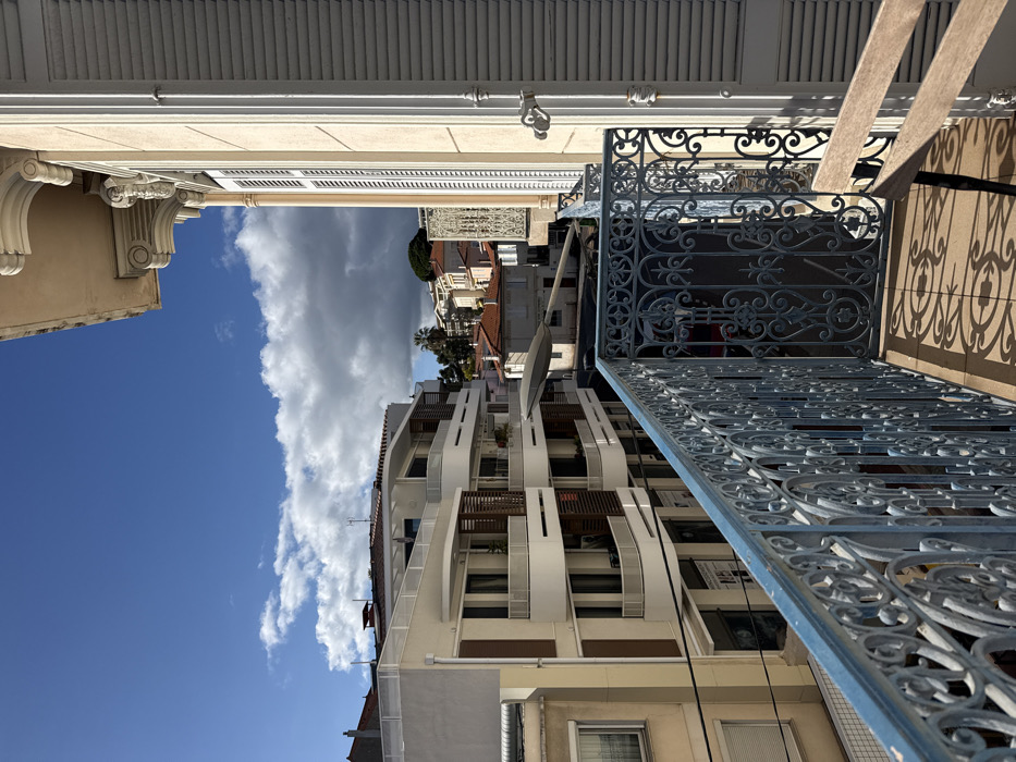 The image depicts a daytime view from a balcony in Cannes, France, looking outward into a residential and commercial cityscape. In the immediate foreground, a weathered light-blue wrought iron balcony railing with elaborate scrollwork patterns extends horizontally across the frame. Distinct shadows from this railing are cast onto the light-colored tiled balcony floor, where a portion of a light-brown wooden chair is visible in the lower right. To the right of the balcony, a segment of a light-beige building facade with classical architectural embellishments, including decorative cornices and an open white louvered shutter, is present.
Across from the viewpoint, a multi-story, modern building with light-colored facades and multiple tiered balconies featuring white railings and dark brown vertical slatted privacy screens is prominent. Below this building, storefronts with glass windows are visible at street level. A light-colored street lamp or utility pole extends diagonally from the lower left towards the upper right midground, passing between the buildings. In the background, further buildings with traditional red-tiled roofs are nestled among greenery, including palm trees, suggesting a hilly terrain. The sky is partially clear and blue with scattered white cumulus clouds, while a darker, denser cloud mass is visible on the right side of the frame. No individuals are visible.