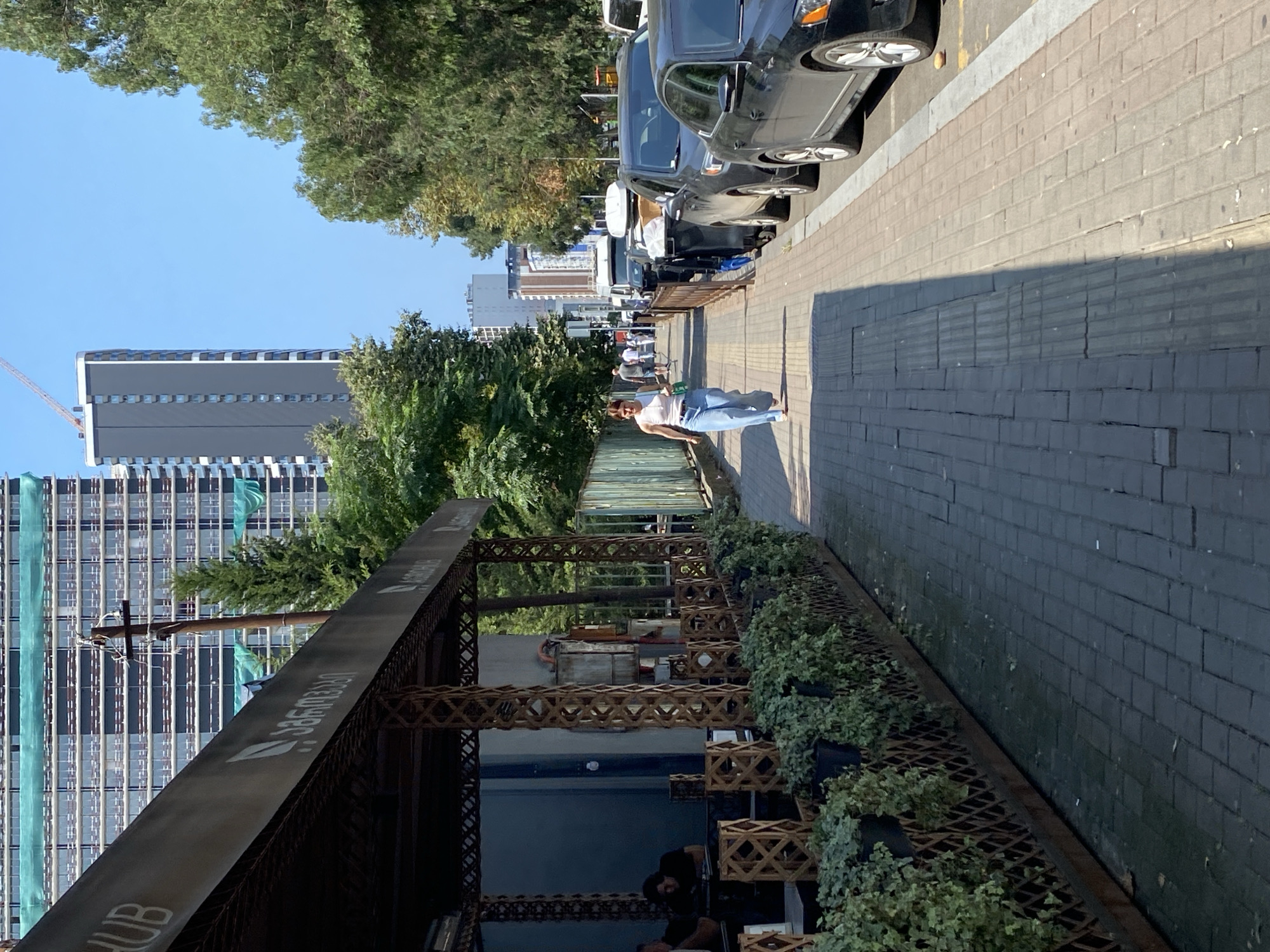 A woman is walking on a sidewalk in Batumi, Georgia. She is wearing a white tank top and blue jeans. The sidewalk is made of brick and there are trees on the left side of the sidewalk. On the right side of the sidewalk, there are cars parked on the street. In the background, there are tall buildings. One of the buildings appears to be under construction. The weather is sunny and clear.