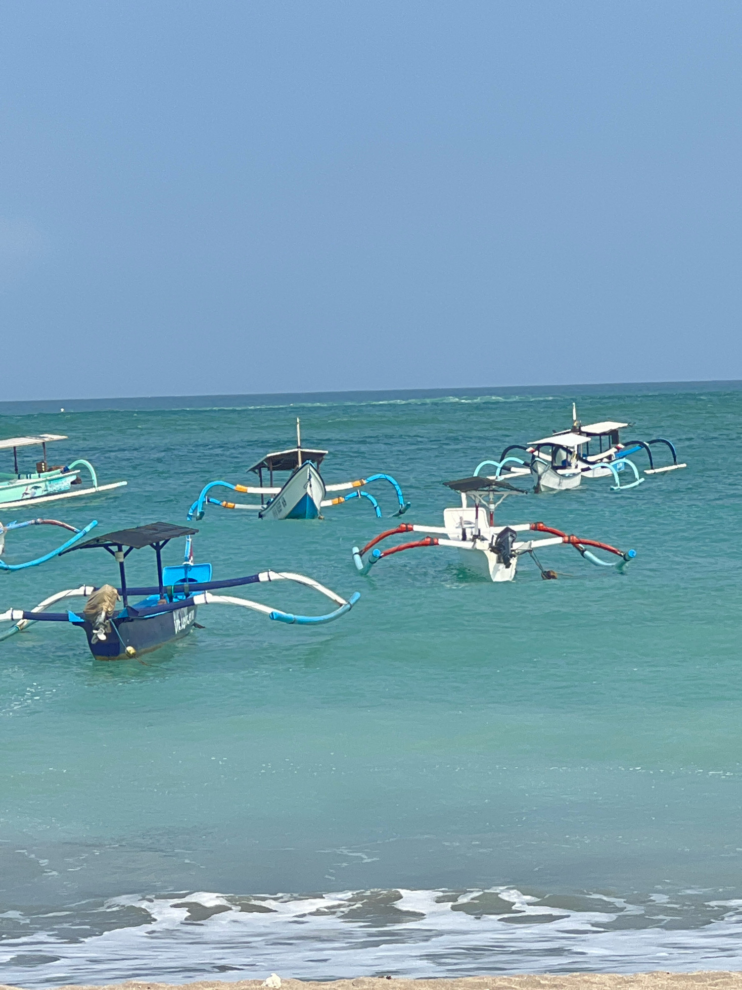 The image shows a calm, turquoise ocean with several traditional Balinese fishing boats, known as "Jukung," anchored offshore. The boats are characterized by their outriggers, which extend on either side to provide stability. The sky is a clear, pale blue, indicating a sunny day. The water is clear enough to see the sandy bottom in some areas. In the foreground, gentle waves are lapping at a sandy beach. The scene is characteristic of the Kuta coastline in Bali, Indonesia, a popular tourist destination known for its beaches and watersports. No people are visible in the image, suggesting the boats are either temporarily unattended or waiting for passengers. The overall impression is one of peacefulness and tranquility.