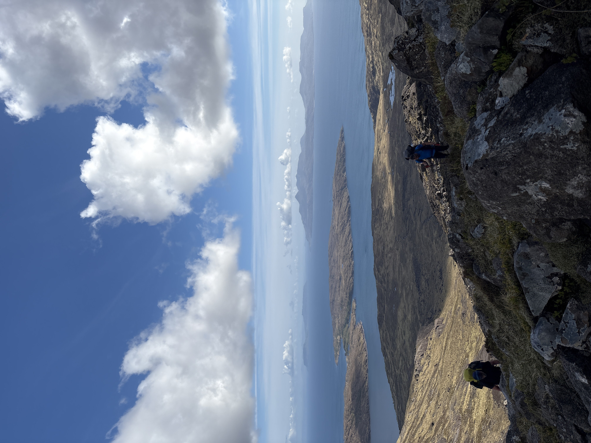 The image shows a landscape scene with a prominent rocky outcrop in the foreground, a wide expanse of water with islands in the middle distance, and a sky filled with clouds. Two figures are visible, seemingly hiking on the edge of the rocky outcrop. The environment suggests a mountainous or coastal terrain, likely a part of the Isle of Skye in the United Kingdom. The weather appears clear with sunny conditions allowing good visibility across the scene.