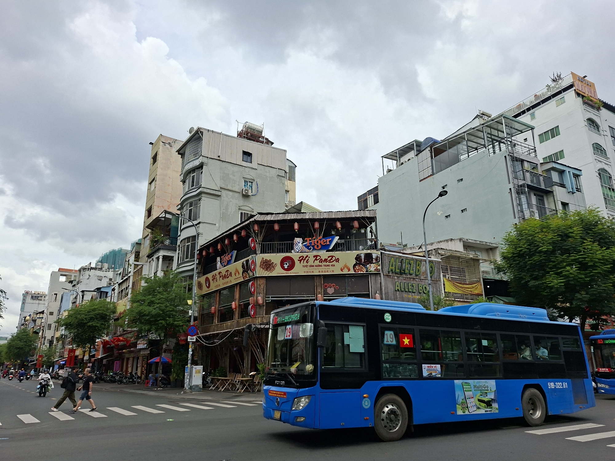 The image captures a street scene in Thủ Đức, Vietnam, on a cloudy day. A bright blue bus with the number "19" prominently displayed is in the foreground, traveling on a paved road with a pedestrian crossing. The bus features advertisements on its side, including one for "MultiGo." On the right side of the bus, a Vietnamese flag is visible.
The street is lined with multi-story buildings, many of which house businesses, indicated by signage. One building features a sign for "Tiger Beer" and "Hi PanDa Chinese BBQ & Hotpot." Another sign reads "ALLEZ BO." Trees and awnings are visible along the sidewalks.
Several people are on the street. Two individuals are crossing the pedestrian crossing in the foreground, with one person wearing a backpack. Other people are seen on motorcycles in the background, typical of urban traffic in Vietnam. The architecture suggests a mix of older and more modern structures. The overall atmosphere is that of a bustling city environment.
