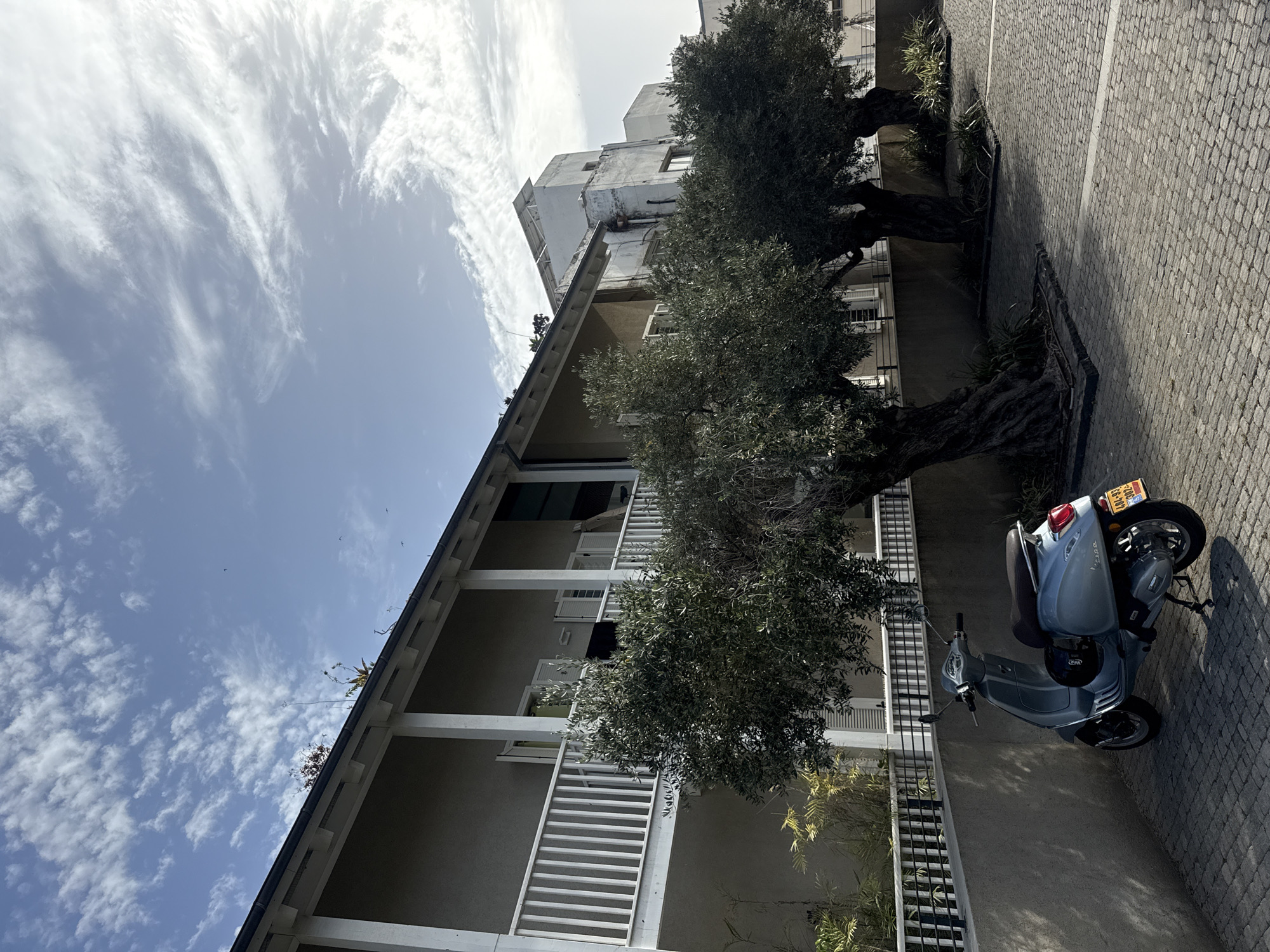 The image depicts a street scene in Tel-Aviv, Israel. The view is looking up at a multi-story building with white railings and balconies, partially obscured by mature trees. A light blue Vespa scooter is parked on the cobblestone pavement in front of the building. The sky is visible above the building, featuring light clouds.