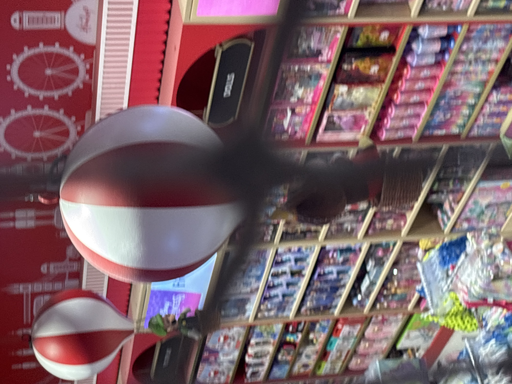 The image depicts a toy store interior. Two large red and white striped balloons are prominently displayed, suspended from the ceiling. In the foreground, a dark, blurred object partially obscures the view of the balloons and the merchandise below.
To the left of the larger balloon, a small figurine of the character Luigi, from Nintendo's Super Mario franchise, is positioned on a shelf. Behind the balloons, the store's background features a red wall with white graphics of Ferris wheels and cylindrical structures, possibly representing landmarks or a themed design.
Below the balloons, shelves are filled with various toys and products. Some shelves hold packaged dolls and action figures, while others display rows of colorful bottles and containers. A section of the store appears to be dedicated to dolls, as indicated by a black sign with the word "DOLLS" in white lettering. The overall impression is a brightly lit, well-stocked retail environment.
This scene is located in a toy store in Dubai, United Arab Emirates, likely within a shopping mall.