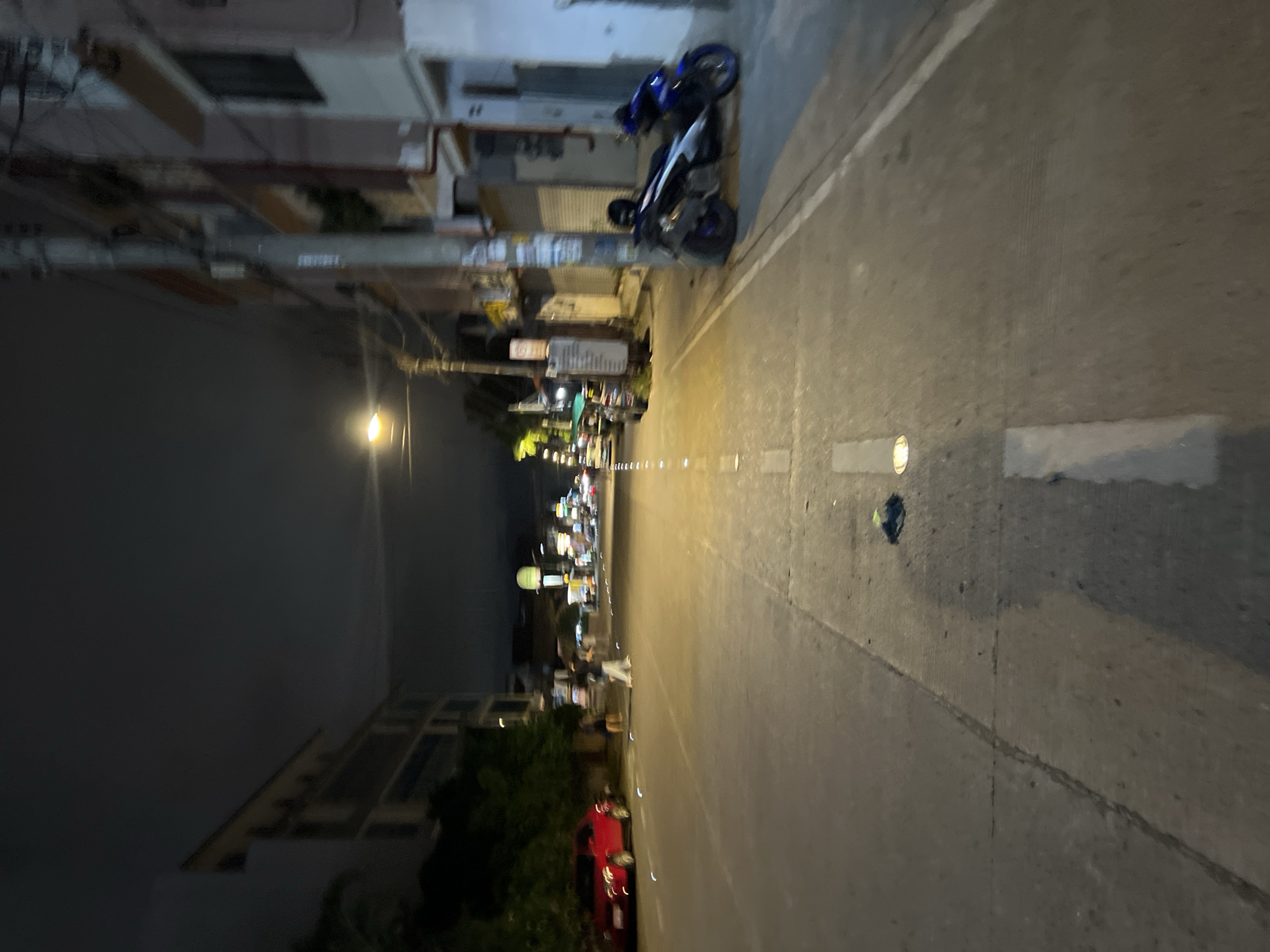 The image depicts a street scene at night in Davao City, Philippines. The street is paved with concrete and marked with white lines. On the left side of the street, a red car is parked, and further down, there are trees and buildings. Several people are walking on the street. On the right side of the street, there are buildings, a utility pole, and two motorcycles parked. The sky is dark, indicating nighttime. Streetlights illuminate the scene.