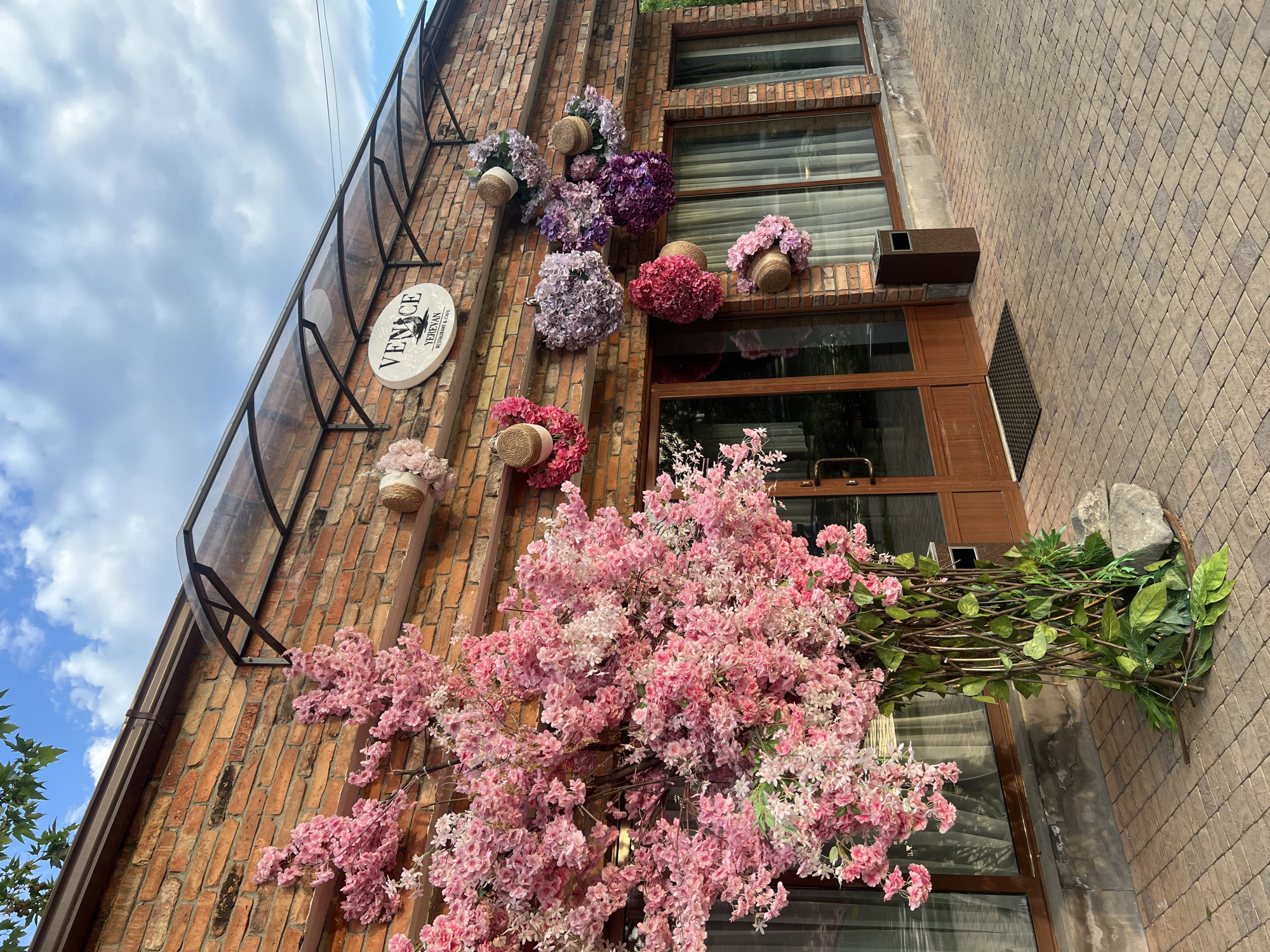 The image depicts the exterior of a restaurant and cafe named "VENICE YEREVAN". The building is constructed with red brick and features a transparent awning above the entrance. Large windows with sheer curtains are visible.
In front of the building, a large, artificial tree adorned with pink cherry blossoms stands prominently. The base of the tree is wrapped in woven branches and greenery, with stones placed around it. The entrance to the establishment is marked by wooden doors and a black doormat.
On the brick wall, above the windows, there are several decorative arrangements. These include a circular sign for "VENICE YEREVAN RESTAURANT & CAFE", and multiple round floral arrangements in various shades of pink, purple, and lavender. Some of these floral decorations are complemented by straw hats.
To the right of the entrance, a dark brown trash receptacle is situated. The ground in front of the building is paved with interlocking beige and brown stones. The sky above is blue with scattered white clouds. The overall scene suggests a pleasant, possibly springtime, outdoor setting for a dining establishment.