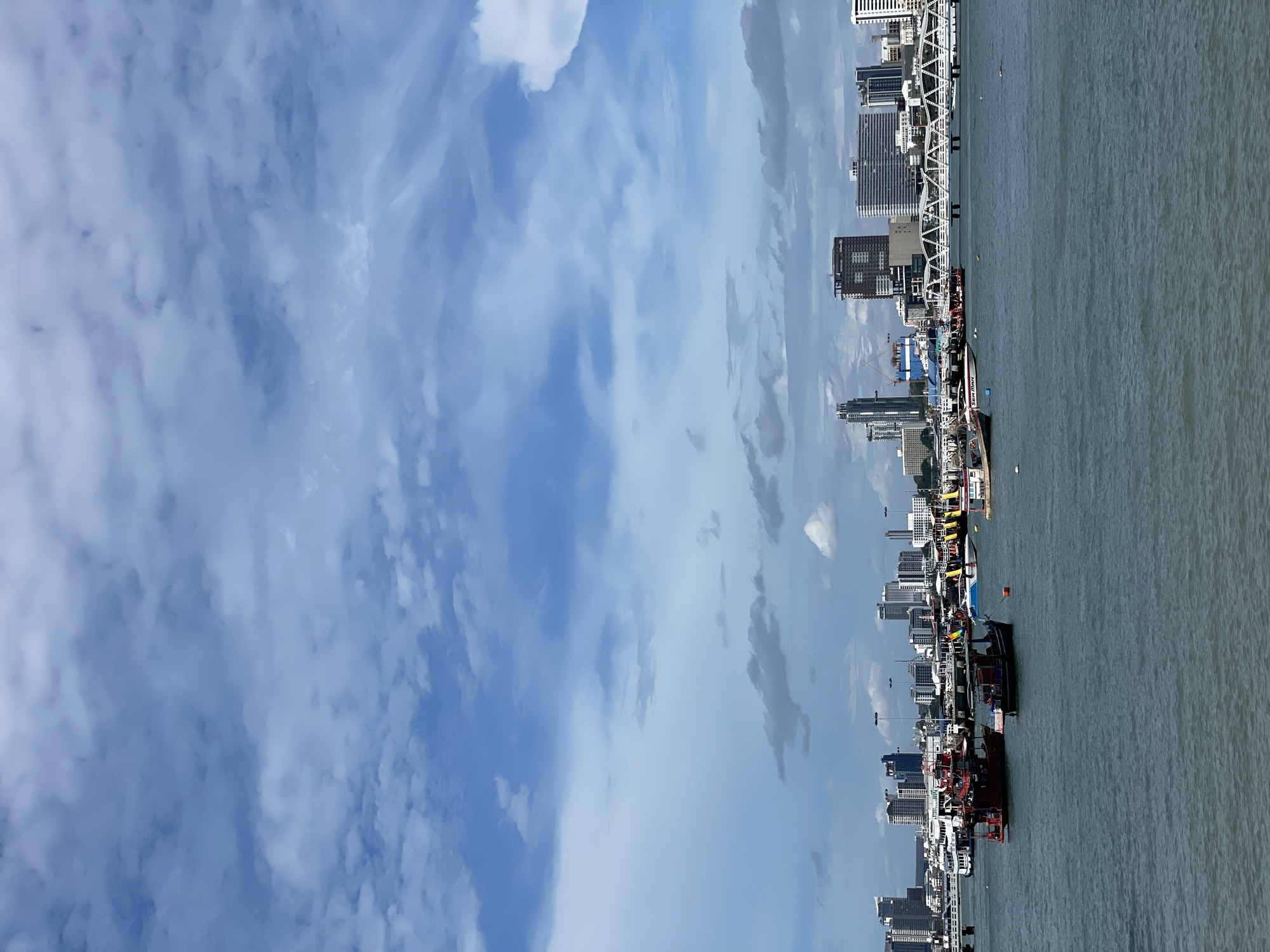 The image shows a wide shot of the Pattaya City coastline in Thailand, captured from the water. The sky is a mix of blue and white, with large, scattered clouds. In the foreground, the sea is a choppy, dark teal. A number of boats are visible, ranging from small fishing vessels to larger passenger boats. Some boats are moored, while others appear to be in motion. The middle ground features a modern cityscape with numerous high-rise buildings and a prominent bridge. The buildings are of various architectural styles and heights, with many appearing to be hotels or condominiums. The bridge spans across the water, connecting different parts of the city. The overall scene depicts a busy urban waterfront area. There are no visible people in the image.