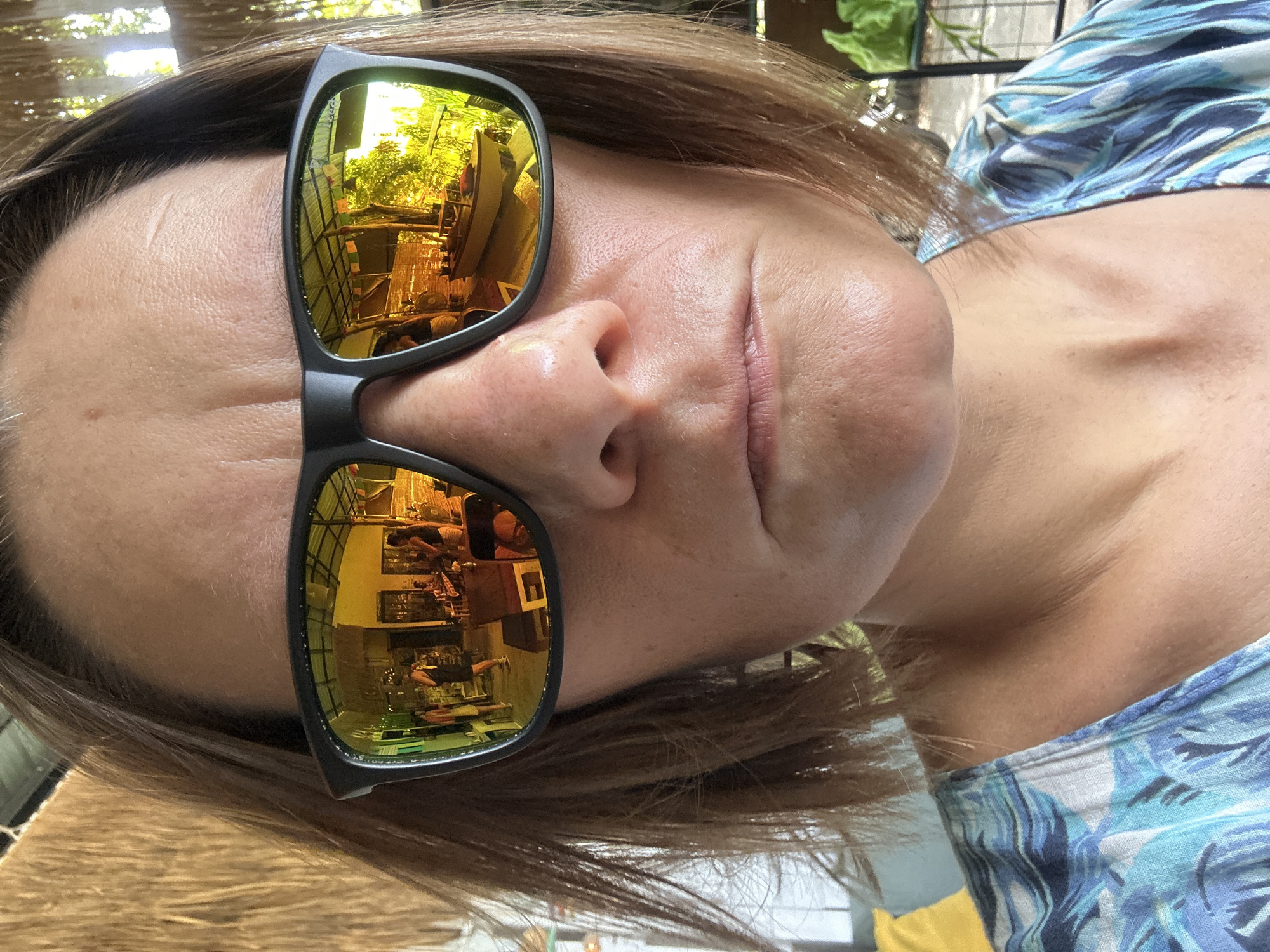 A woman is photographed close-up wearing sunglasses with a reflective gold lens. The reflection shows an indoor restaurant scene. She has fair skin, brown hair, and is wearing a blue patterned shirt. The setting appears to be ตำบลเกาะพะงัน, Thailand, based on the probable location of the restaurant reflected in the sunglasses.
