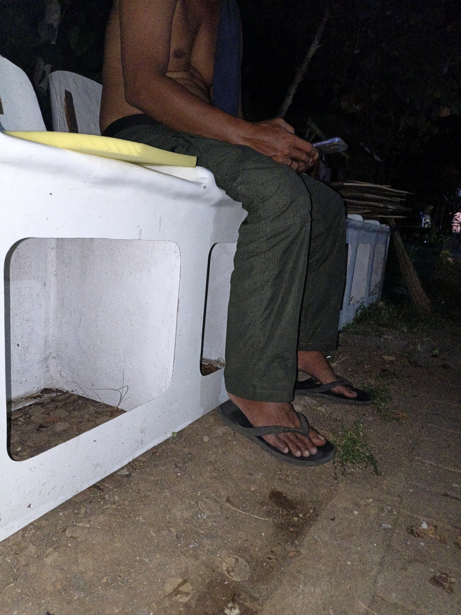 A person sits on a white concrete bench outdoors at night, holding a mobile phone. The person is wearing dark green pants and dark flip-flops, and their upper body is bare. The bench has two open rectangular sections. The ground is dirt and gravel, with some sparse vegetation. In the background, there are dark trees and a pile of wood. The overall scene appears to be in Cikal, Indonesia.