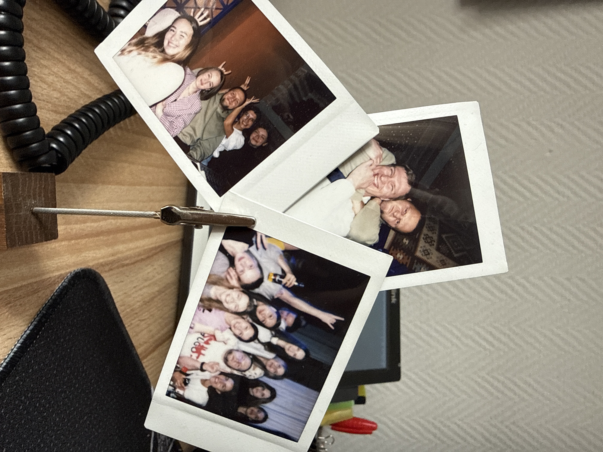 The image shows three Polaroid photographs clipped to a stand on a wooden surface, with a coiled black cable and black textured object nearby. The photographs appear to depict groups of people in various indoor settings. The top photograph shows a group of young women and men posing for the camera. The second photograph shows two men embracing and smiling. The third photograph features a larger group of young women posing together. The location cannot be determined from the image.