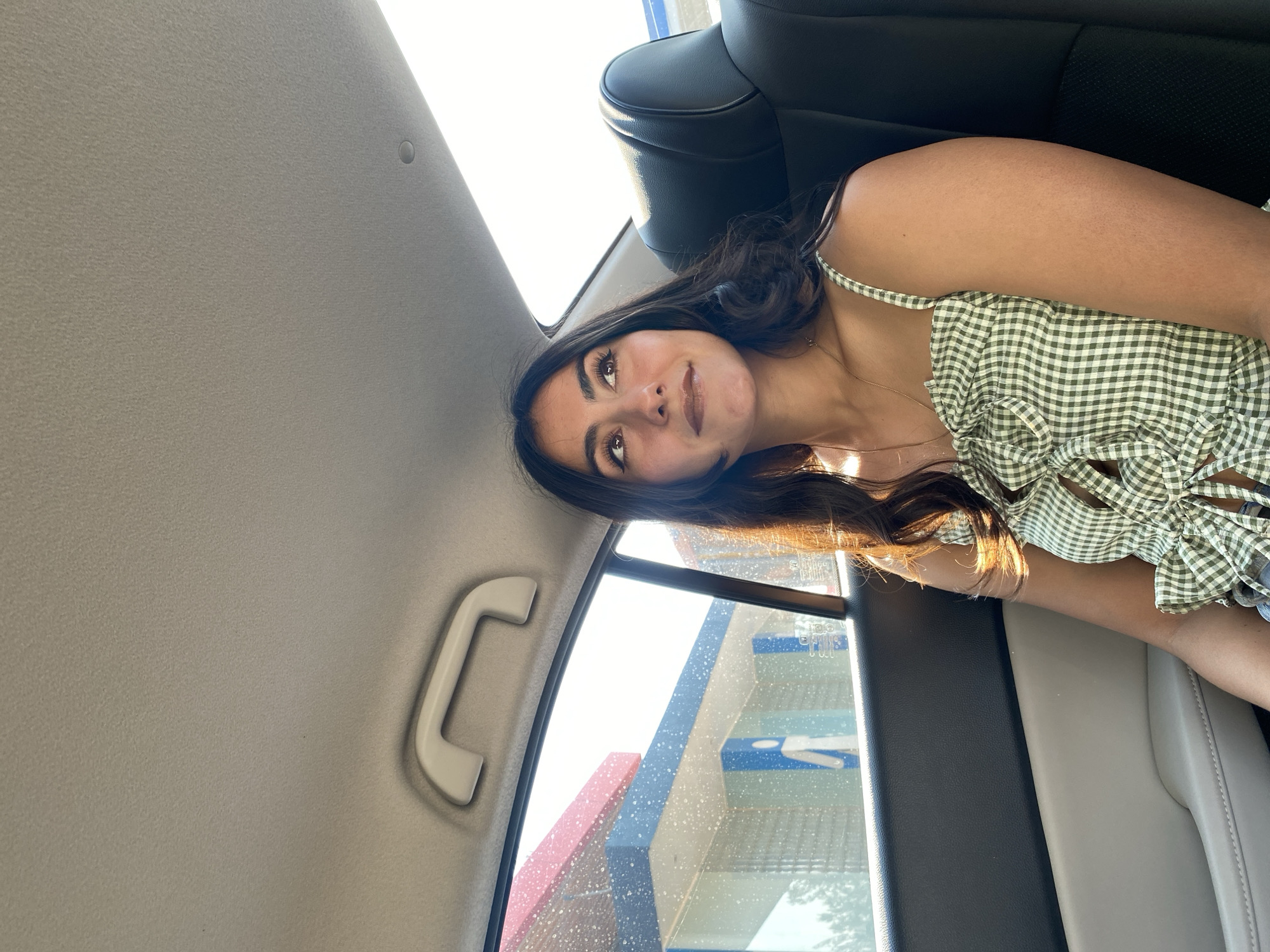 A medium shot captures a young woman in the back seat of a car. She is wearing a green and white gingham top with spaghetti straps. Her long, dark hair cascades around her shoulders. Her gaze is directed upwards and slightly to the left. The car's interior is visible, including the seat, headrest, and ceiling. Outside the window, abstract shapes and colors are visible. Based on the color scheme and shape of the building it looks like the building is a Wal-Mart. Santa Ana Xalmimilulco, Mexico, is not visible within the image, it is not possible to determine if the image was taken there based on the available data.