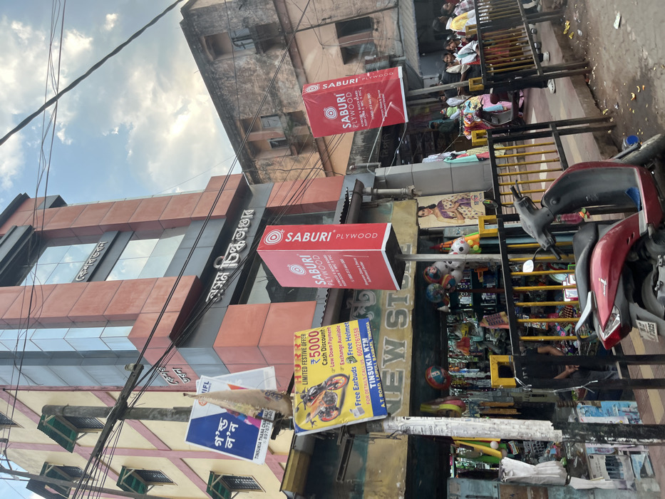 This image captures a bustling commercial street scene in Tinsukia, India, under a partly cloudy daytime sky. The perspective is tilted, looking upwards at several buildings and street-level activities.
The main building in the center is a multi-story structure with reddish-brown cladding and reflective glass windows. A large sign in Bengali/Assamese on this building reads "অসম চিল্ক ভাণ্ডার" (Assam Silk Bhandar), indicating a silk store. Below it, two prominent red rectangular signs, hanging perpendicularly from the building, advertise "SABURI PLYWOOD," complete with contact details.
At street level, various shops are visible. One storefront displays a wide array of toys and household goods, while another appears to be a clothing store with garments hanging on racks. Several people, including children, are seen on the sidewalk, some browsing merchandise, others standing or sitting. A red and black scooter is parked on the right, near metal railings that separate the pedestrian area from the road.
Numerous banners and signs hang above the street. A large yellow and blue banner advertises "TINSUKIA KTM," promoting a "LIMITED FESTIVE OFFER" that includes a "₹5000 Cash Discount," "Low Down Payment," "Exchange Available," "Free Earbuds," and a "Free Helmet." It also provides a contact number (6002761558) and address: "A.T. ROAD LAIPULL BANBARL TINSUKIA." Another smaller blue banner advertises "গ'ল্ড ল'ন" (Gold Loan), featuring a phone number and mentioning "TINSUKIA THANA CHA." An "IFL" sign is also visible. A dense network of overhead power lines crisscrosses the upper portion of the frame. The scene conveys the vibrant atmosphere of a busy commercial area.