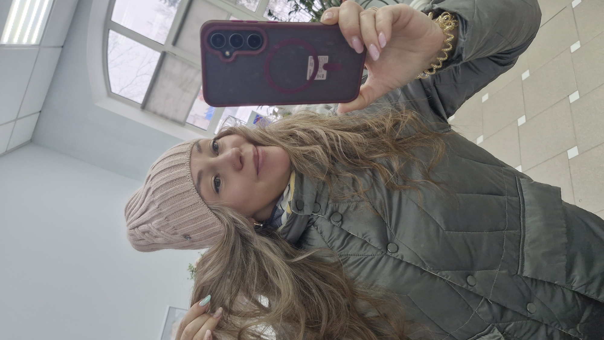 A woman takes a selfie indoors in Otchor, Russia. She holds a dark red/purple smartphone, which features a triple camera setup and a ring stand on its back, capturing her reflection. She has long, wavy, light brown and blonde hair, which cascades over her shoulders and jacket. She wears a light pink knitted beanie, a dark green quilted jacket, and a blue and white checkered scarf peeking out. A gold-colored coiled bracelet adorns her right wrist, and her fingernails display alternating light pink and light blue/green polish. A subtle nose piercing is also visible.
Her expression is serene, with a slight smile as she looks directly into the camera. The background indicates a public or commercial indoor space, characterized by light blue/grey walls, a high ceiling, and rectangular overhead lighting fixtures. An arched window reveals a view of a grey sky and bare tree branches outside, suggesting a cold, overcast day. The floor is tiled with light-colored squares interspersed with smaller white squares. A small, illegible white sticker is present on the back of her phone case.