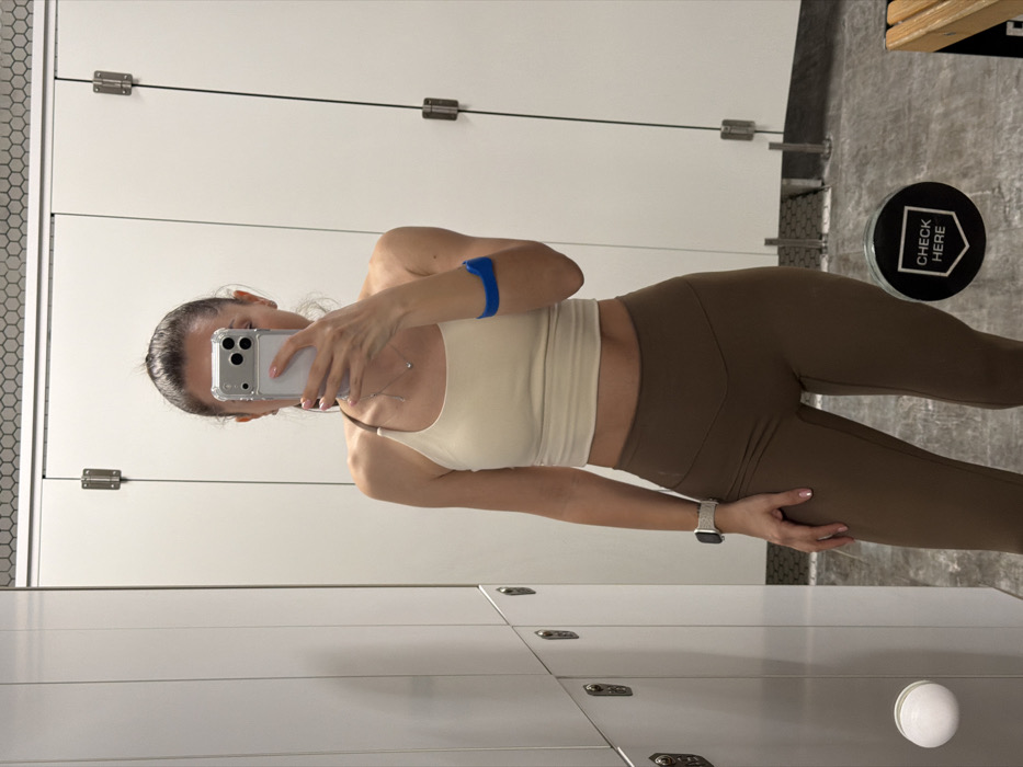 The scene depicts an interior view, likely a changing or locker room located in Varna, Bulgaria. A female individual, positioned centrally, is reflected in a mirror, holding a silver-colored smartphone with a clear case in her right hand, obscuring her face to capture a self-portrait. She has dark hair pulled back. Her attire consists of a cream-colored asymmetrical crop top and dark brown high-waisted leggings. A light-colored smartwatch with a textured band is present on her left wrist, and a blue athletic band is worn on her right upper arm. A delicate silver chain with a small pendant is visible around her neck. The background features a row of white locker doors with silver-toned locks and white circular knobs to the left. To the right, two white stall doors, likely for changing, are visible with their hinges. Above these doors, a strip of small white hexagonal tiles lines the upper wall. The floor is dark and appears to be concrete or a similar material. In the lower right corner, a circular black sign with white text reading "CHECK HERE" is reflected on the floor, next to a segment of a light-colored wooden bench.