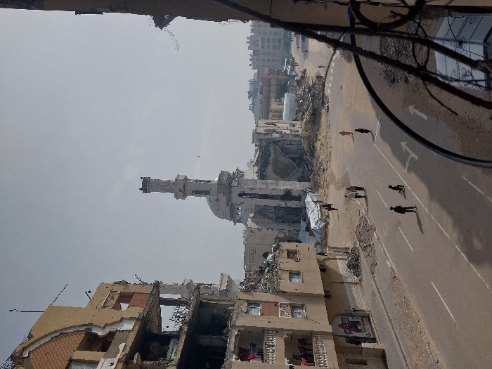 An elevated view captures a heavily damaged urban street scene in Khan Yunis, Palestinian Territory, under a hazy sky. To the left, a multi-story, light brown building exhibits extensive structural damage, with upper floors largely collapsed, revealing exposed rebar and interior spaces. A group of individuals, primarily women in red garments, are visible on a lower balcony of this building. Debris is piled along its base on the sidewalk.
In the center of the frame, a tall, white mosque minaret stands, showing signs of damage but largely intact, while the mosque's main building behind it is significantly destroyed, with its dome partially visible amidst large quantities of rubble. Widespread demolition debris and mounds of dirt occupy the areas surrounding the mosque and adjacent to the street, indicating complete structural collapse.
The asphalt road below is marked with white lane lines, including two forward-pointing arrows. Several individuals navigate this road: two adults walk together away from the camera, while further down the street, two solitary figures walk in the same direction. Closer to the foreground, two individuals, appearing to be children, are positioned near the road's center line; one stands upright, and the other kneels or crouches on the asphalt.
To the right, the immediate foreground is partially obscured by a dark metal railing. In the mid-distance on the right, temporary shelters constructed with white tarpaulins are visible amidst further rubble and debris. More distant buildings, some appearing less damaged, define the city horizon.