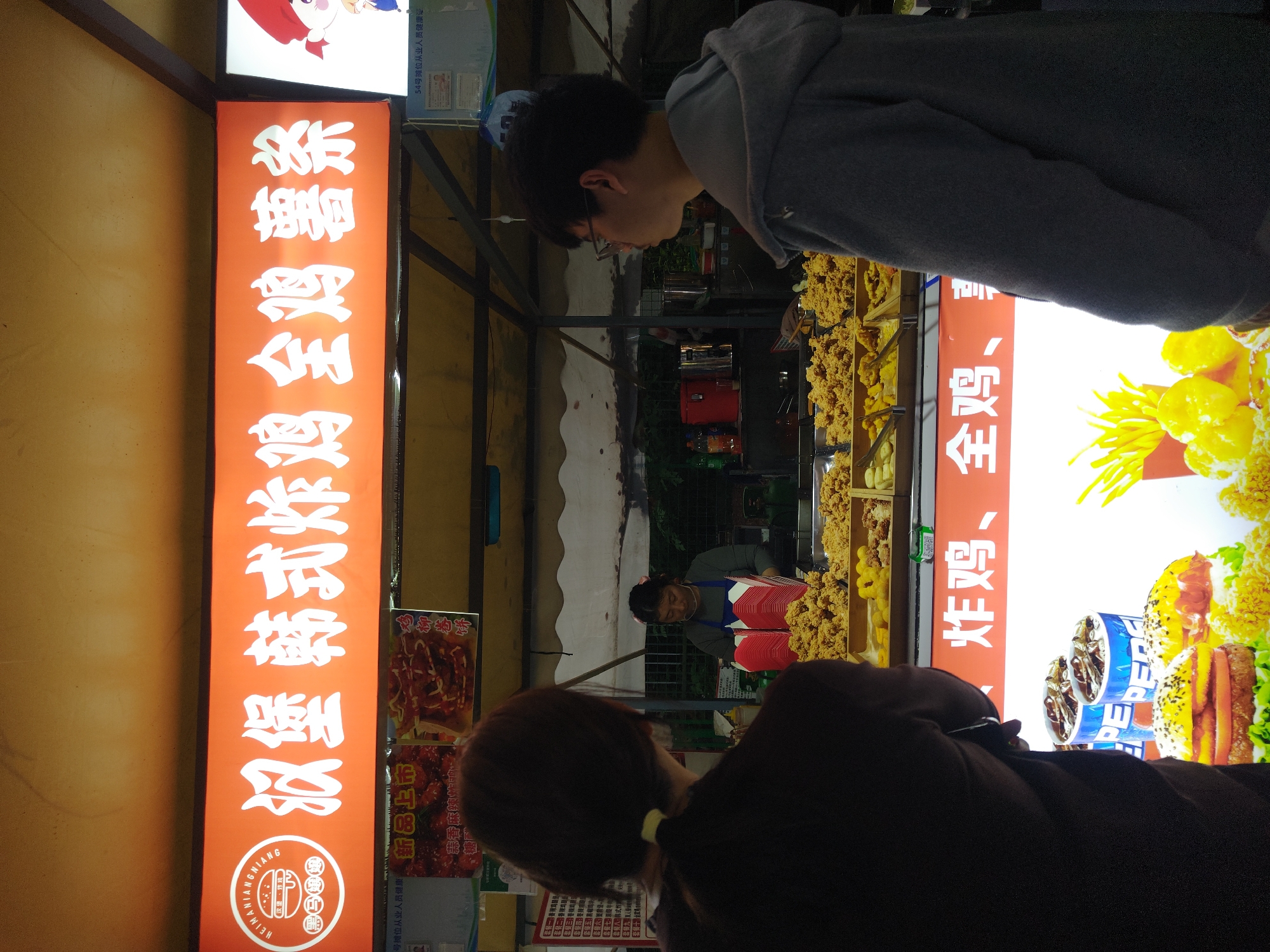 The image shows a street food stall in Shapingba District, China. A person wearing glasses and a grey hoodie is looking at the food displayed. Another person with dark hair is also looking at the display. The stall worker is visible behind the food display. The stall sells fried chicken and other fried items. Orange signs with white Chinese characters advertise the food. A Pepsi advertisement is also present.