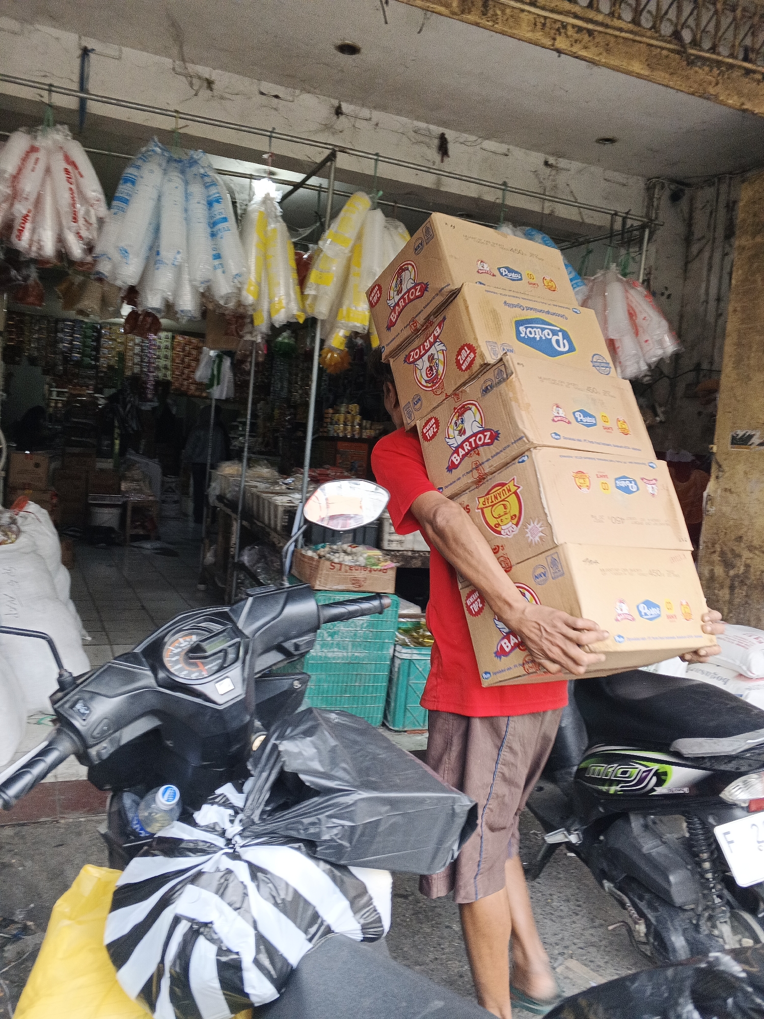 A man wearing a red t-shirt and brown shorts carries a stack of five cardboard boxes, each labeled with various brand names, including "BARTOS" and "Potios". He is walking in front of a market stall in Sukabumi, Indonesia. The stall is stocked with goods, including bags of plastic cups hanging from the ceiling and shelves filled with packaged food items. A black motorcycle is parked in the foreground, loaded with plastic bags. The overall scene depicts a busy marketplace.