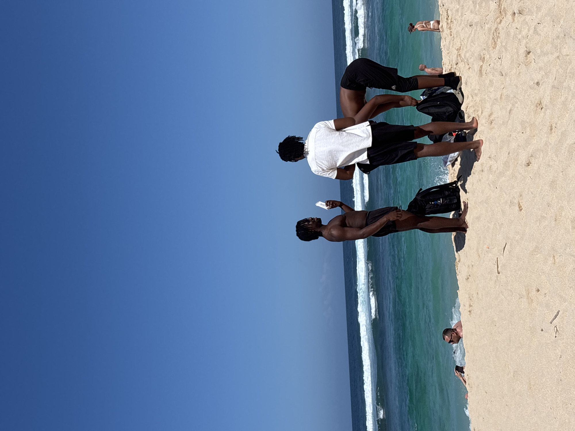 The image depicts a sunny day at a beach, likely Kuta Selatan, Indonesia, as indicated by the context. The vast expanse of a clear blue sky dominates the left side of the frame, suggesting fair weather. The right side showcases a sandy beach and the turquoise waters of the ocean, with visible waves breaking near the shore.
Several people are present on the beach. In the foreground, three young men are visible. One is standing and looking down, holding a white object, possibly a phone, in his hand. Another man, wearing a white t-shirt and black shorts, is also looking down. A third man, in black shorts, is positioned behind them, in a bent-over or squatting posture. Further up the beach, a person in white swimwear is lying on the sand. Near the water's edge, another person is partially visible, appearing to be wading in the ocean.
The overall atmosphere seems relaxed and casual, consistent with a beach outing on a bright day. The light suggests it's midday or early afternoon. There are no discernible text elements in the image. The scene captures a moment of leisure and recreation on a tropical beach.