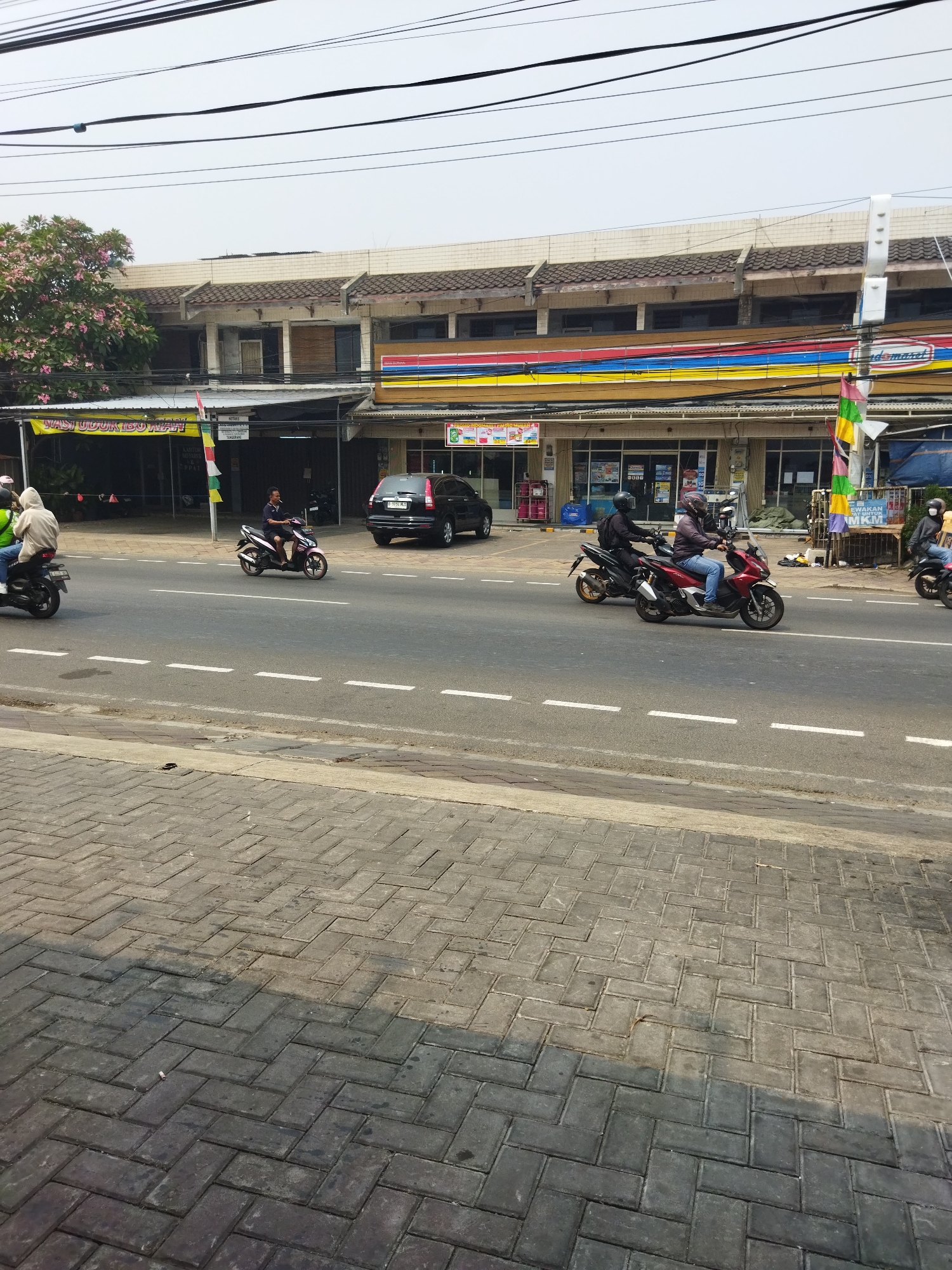 The image depicts a street scene in Tangerang, Indonesia, with a focus on a row of shops and several motorcycles navigating the road. In the foreground, a sidewalk paved with interlocking bricks is visible, with a portion shaded. The road itself is asphalt with white dashed lines indicating lanes.
Several motorcycles are in motion. On the left, a person in a light-colored hoodie rides a black motorcycle. Further down the road, a person in a dark helmet and jacket rides a purple and white motorcycle. To their right, another person in a dark helmet and jacket rides a red motorcycle. In the background, a black SUV is parked on the side of the road.
Behind the road, a strip of shops lines the street. One shop has a prominent sign with red, yellow, and blue stripes, and a logo that appears to be "Indomaret". Other shops have awnings and display signs in Indonesian. A tree with pink flowers is situated to the far left of the shops. Overhead, a network of power lines crisscrosses the sky. The overall lighting suggests it is daytime, likely late morning or early afternoon, given the shadows.