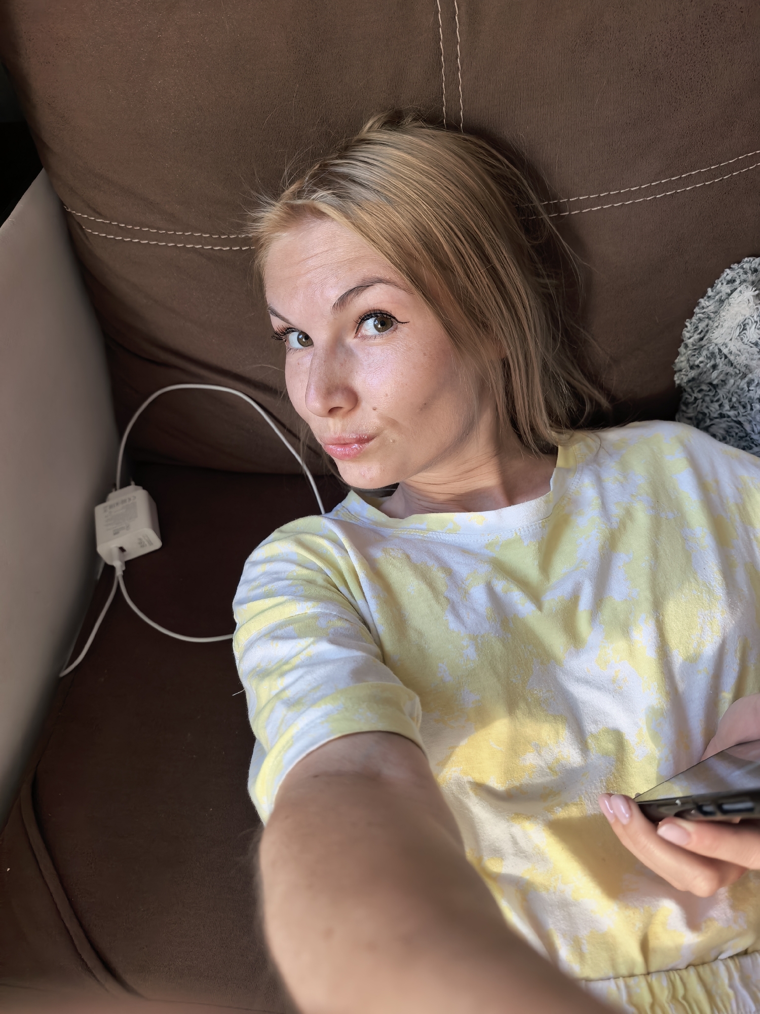 A young woman with light brown and blonde hair is lying on a brown couch. She is wearing a yellow and white tie-dye shirt. She is holding a black smartphone in her right hand. A white phone charger and cable are visible on the couch beside her. The scene appears to be indoors. The location in Sterlitamak, Russia cannot be determined from the image.