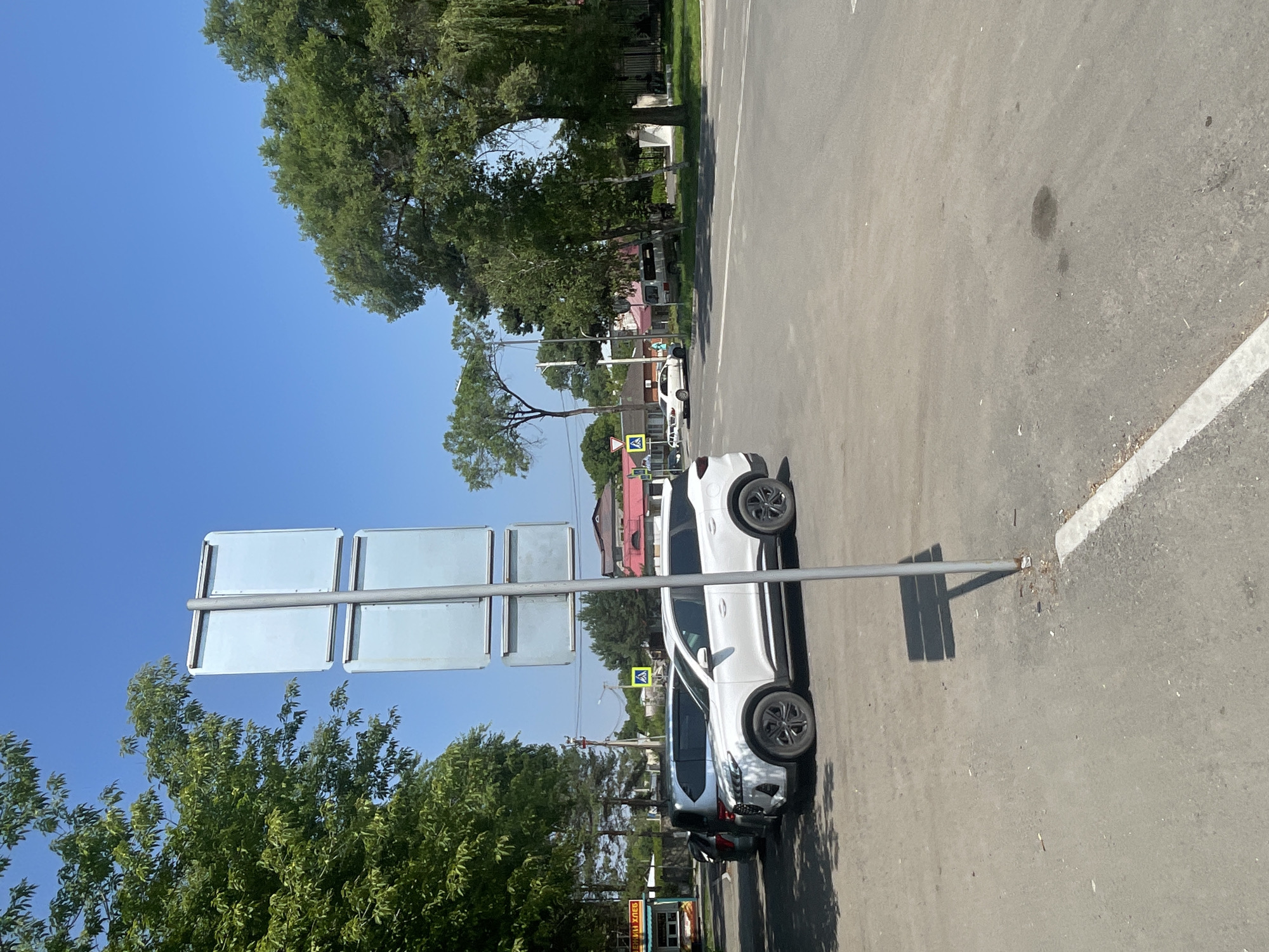 The image depicts an outdoor scene in Spassk-Dal'nij, Russia, under a clear blue sky. In the foreground, a grey metal pole with four blank, light blue rectangular signs is situated on an asphalt surface. The pole casts a square shadow onto the ground. To the left of the pole, a white SUV is parked on a dirt and asphalt area, partially shaded by trees. Behind the white SUV, another vehicle, a silver minivan, is also parked. Further to the left, a small kiosk with green and yellow signage is visible. The background features a street intersection with pedestrian crossing signs. More trees and buildings, including houses with red roofs, are scattered throughout the scene. The overall environment suggests a daytime setting in a town or city.