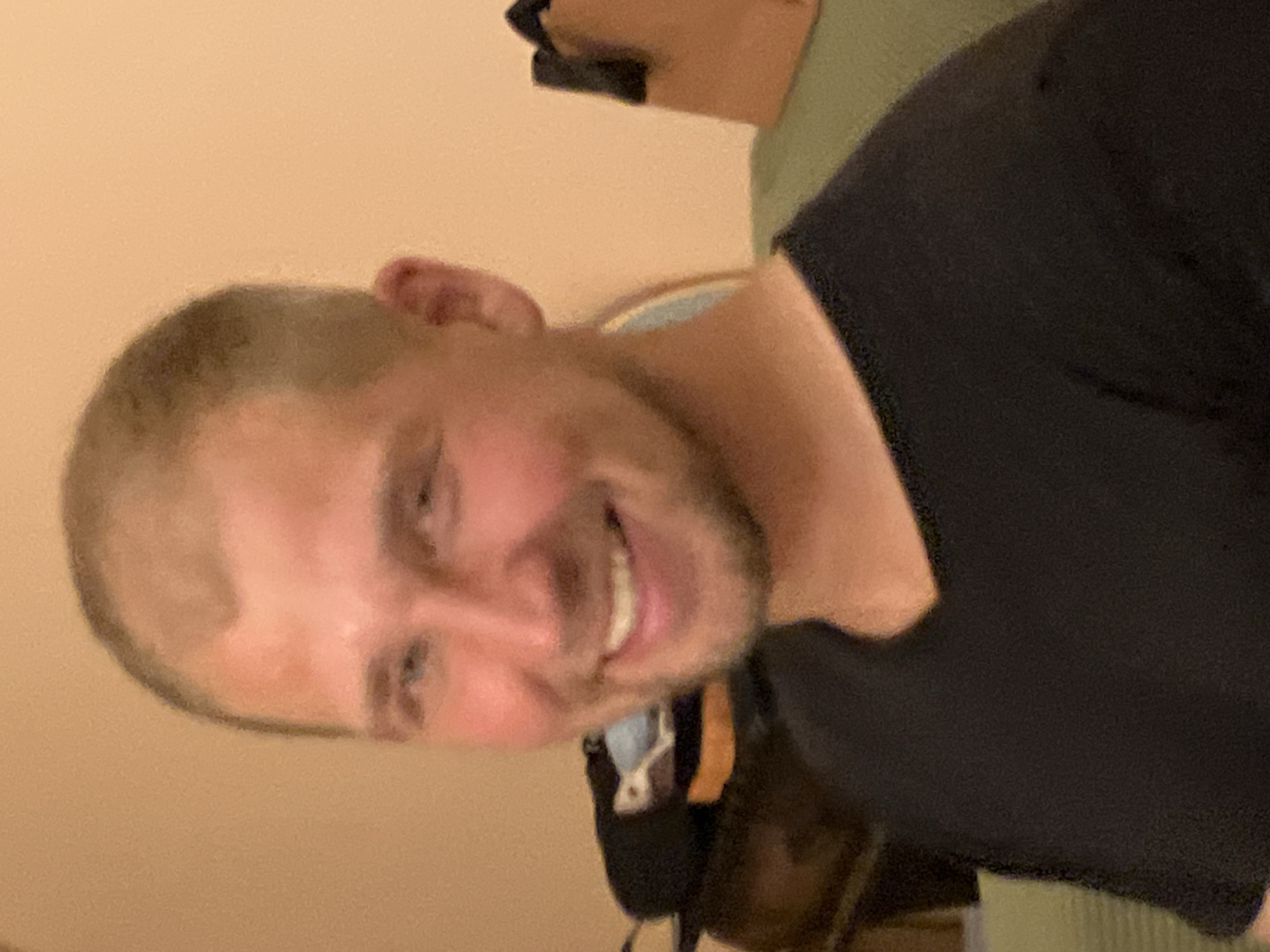 A man with short blonde hair and a stubbled beard is smiling into the camera. He is wearing a black t-shirt. The background is a pale peach wall. The location is not identifiable as being in Tel-Aviv, Israel.
