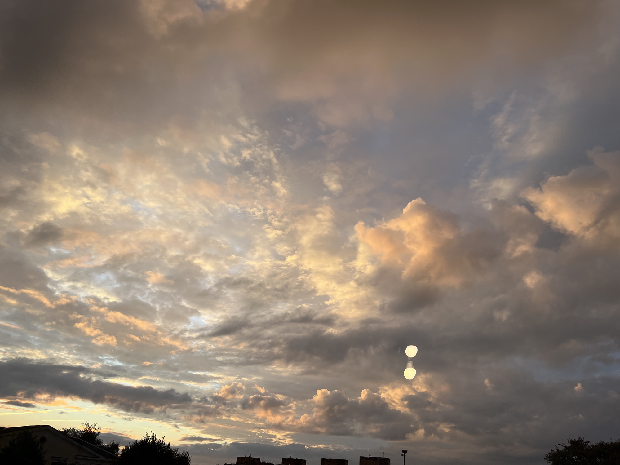 The image captures a dramatic sunset over Kohtla-Järve linn, Estonia. The sky is filled with a dynamic interplay of dark, brooding clouds and lighter, illuminated formations tinged with hues of orange and yellow, suggesting the sun is low on the horizon. In the lower portion of the image, the silhouettes of buildings and trees are visible against the twilight sky. Notably, two bright, oval-shaped lights, possibly lens flares or reflections, are present in the mid-right portion of the frame. The overall scene conveys a sense of atmospheric intensity and the transition from day to night.