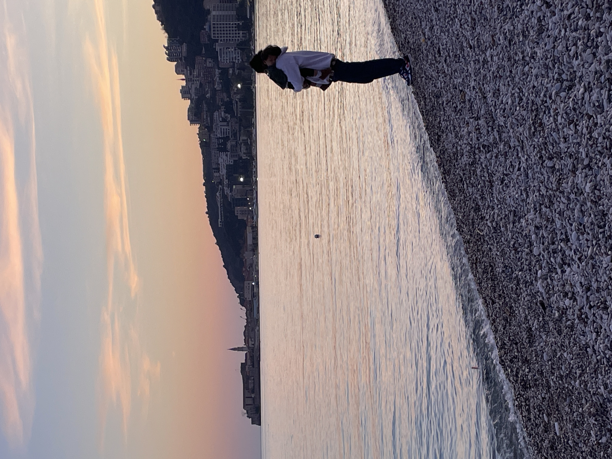 The image depicts a coastal scene at dusk, looking across the sea towards the city of Budva, Montenegro. In the foreground, a gray pebble beach slopes gently towards the water. An adult figure, wearing a light-colored hoodie, dark pants, and athletic shoes, stands facing the water at the edge of the beach, holding a small child who is resting against their shoulder.
The sea occupies the midground, reflecting the twilight sky with calm, undulating surface waves. A small, dark, spherical object, possibly a buoy, is visible floating in the water in the central-left area.
In the background, the historic Old Town of Budva is visible on a promontory to the left, characterized by its fortified structures and a prominent bell tower. To the right, a densely built-up hillside rises, featuring numerous modern buildings with illuminated windows, indicating an urban environment beyond the Old Town. The sky overhead transitions from a soft pink and orange hue near the horizon to a lighter blue, with wispy cloud formations.