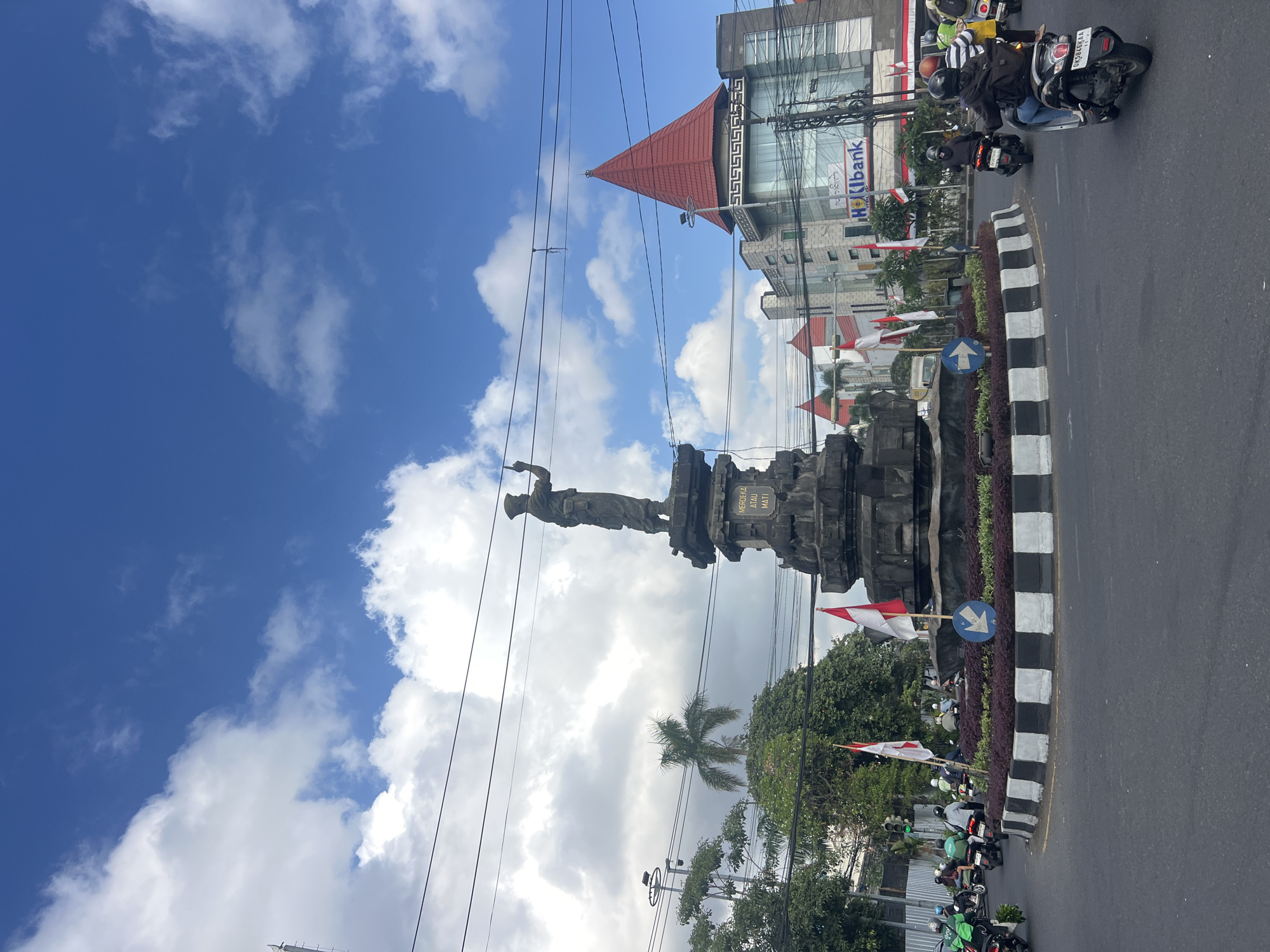 In Denpasar, Indonesia, a traffic circle is depicted with a prominent monument at its center. The monument is a dark-colored stone structure topped with a statue of a standing figure in military attire, holding an upward-pointing object in its right hand. Inscriptions are visible on the monument's base. Surrounding the monument is a circular garden area with dark foliage and white-bordered planters. Several Indonesian flags are planted within the garden.
The traffic circle is active with vehicular traffic, predominantly motorcycles. Several individuals are riding motorcycles, some wearing helmets and backpacks. A building with a distinctive red-tiered roof and glass facade is visible in the background, with signage for "Hokibank". Overhead, electrical wires crisscross the sky, which is a vibrant blue with scattered white clouds. Greenery, including trees and palm trees, frames the scene on the left. Traffic lights are visible in the distance. The overall impression is of a busy urban intersection with a significant historical monument.