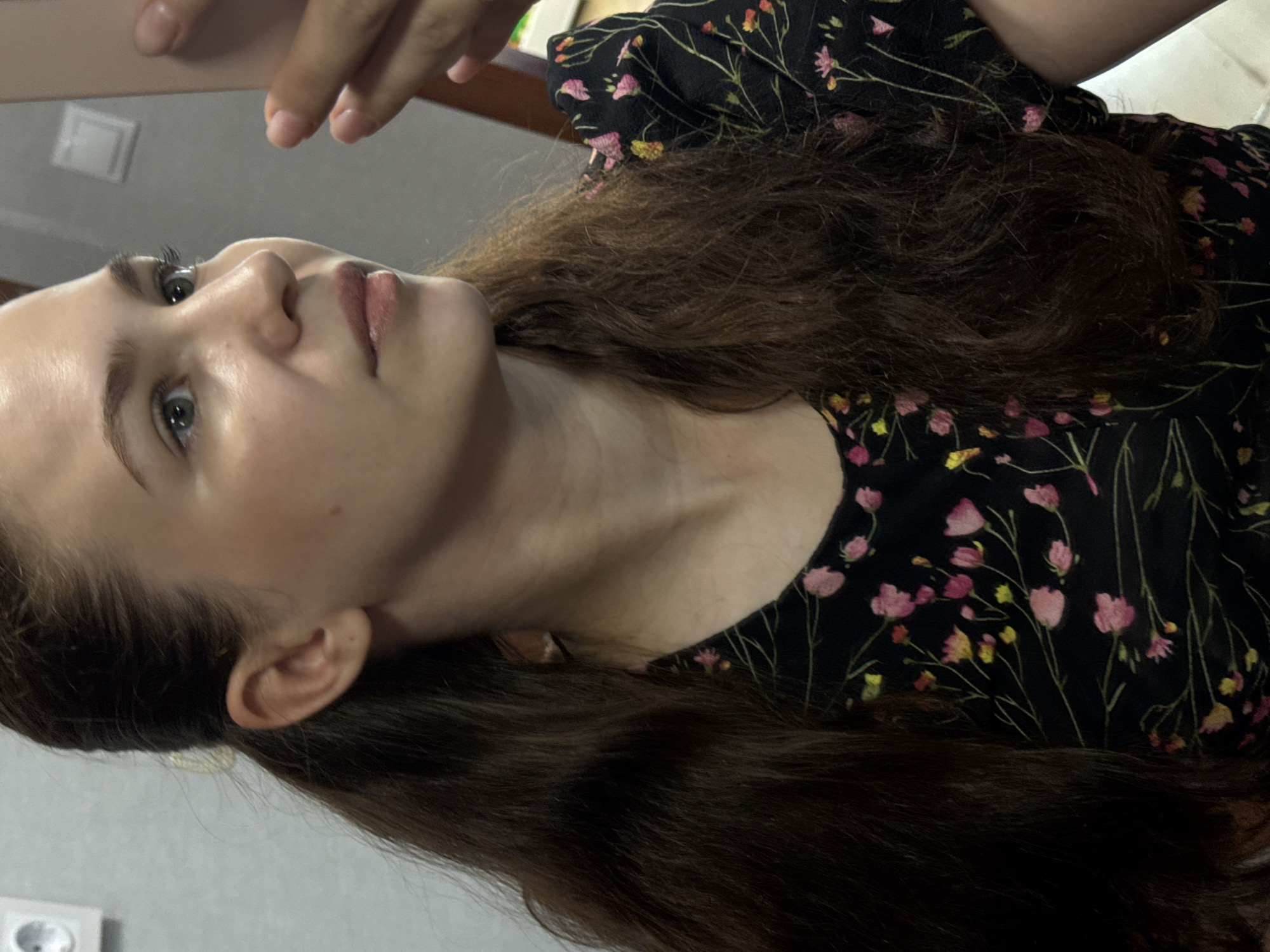 A young woman with long, dark brown hair is shown from the chest up, in a close-up profile shot, looking to her right. She is wearing a black dress with a floral pattern of pink and yellow flowers and green stems. Her hair is partially pulled back with a light-colored clip. She is holding a light-colored object, likely a phone, with her right hand, which is partially obscuring her face. The background is a plain, light-colored wall with a white electrical outlet visible on the left. The lighting suggests an indoor setting. The image does not contain enough information to determine the specific location within Chaikovsky, Russia.