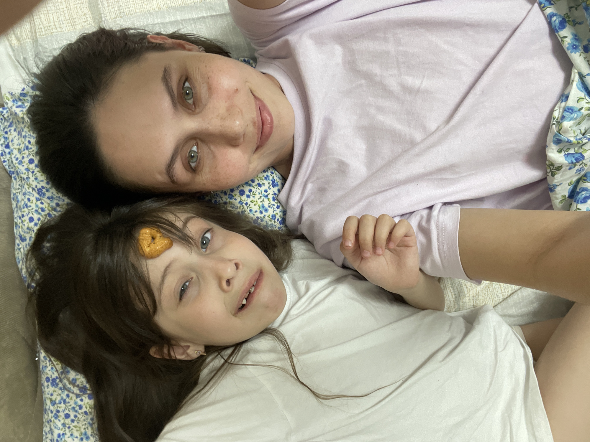 Two people are lying on bedding, one wearing a lavender shirt and the other wearing a white shirt. The younger of the two has a small object on their forehead. The bedding has a floral pattern. Location is unknown and cannot be inferred.
