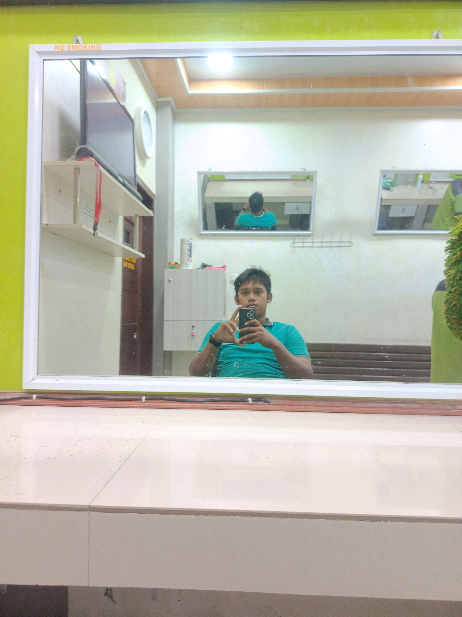 A young man with dark hair, wearing a teal t-shirt, is reflected in a large mirror. He is holding a smartphone in his right hand and appears to be taking a selfie. The mirror is framed by a white border and is mounted on a wall. In the background, there are two smaller mirrors, a clock on the wall, shelves, and some cabinets. The room appears to be well-lit, with light-colored walls and ceiling. There is a "NO SMOKING" sign visible at the top of the mirror frame. The floor in front of the mirror is tiled with a light-colored surface. This scene is set in a business, possibly a barber shop or salon, located in Rijang Pittu, Indonesia.