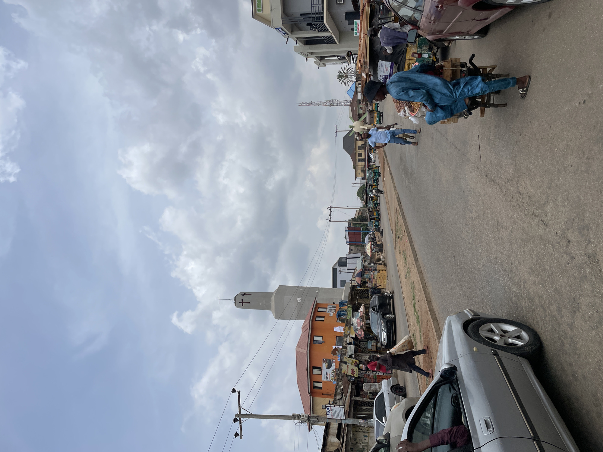 This image depicts a street scene in Jos, Nigeria. A man in a blue outfit and black cap is seen seated on a rudimentary wheeled cart, potentially selling goods. Several cars are parked along the roadside, a church stands prominently in the background, featuring a cross on its tower. Electric poles line the street. Pedestrians are visible amongst the cars and vendors on the side of the road, and market stalls line the side of the road on the right side of the image. The sky is overcast with a heavy cloud cover.