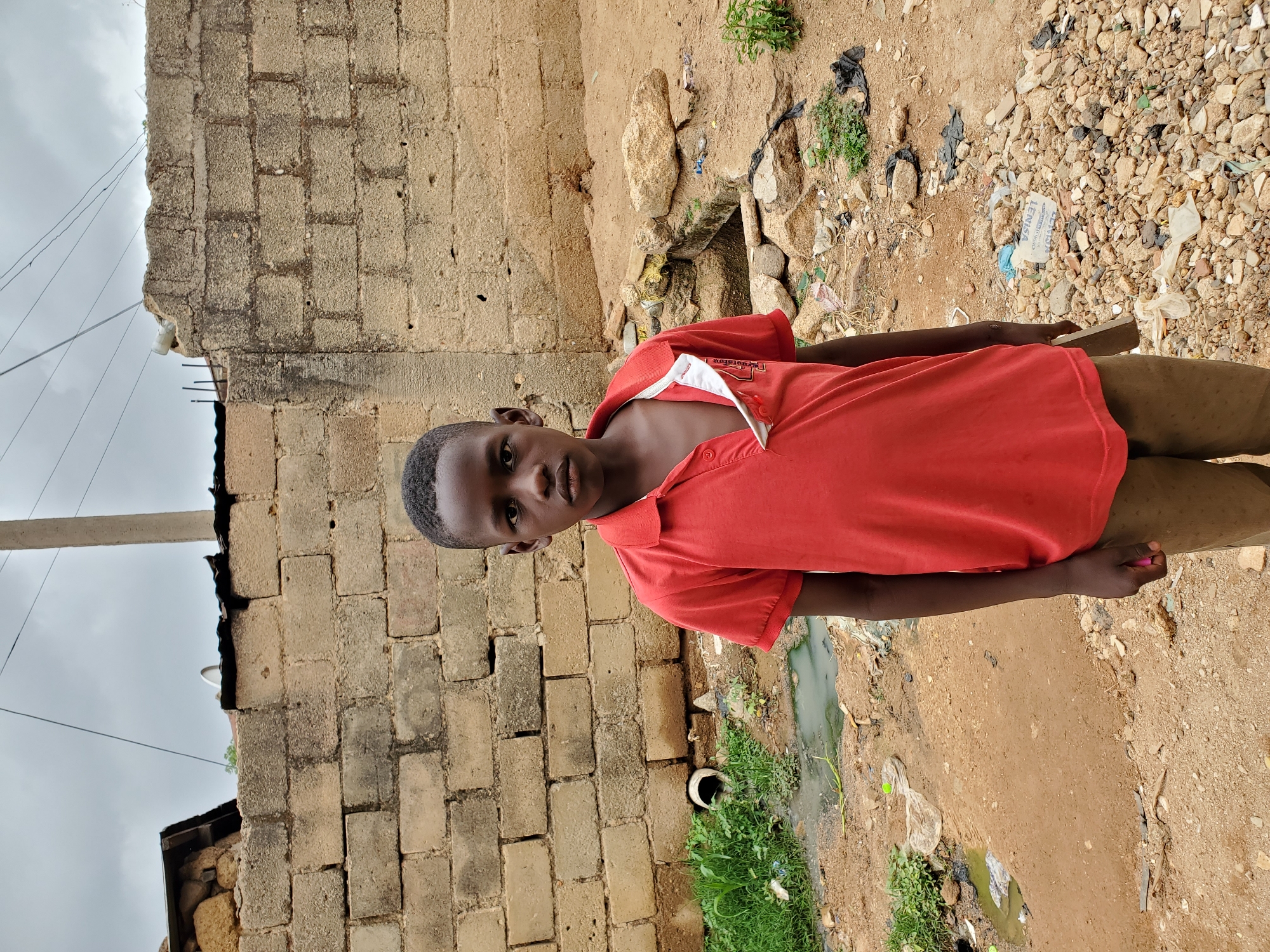 A young boy stands outdoors in Jos, Nigeria, against a backdrop of a concrete block structure and a dirt area with debris. He is wearing a red polo shirt with a white collar and khaki pants. The area surrounding him appears to be a roadside or unpaved area with scattered stones, trash, and vegetation. A pipe is visible along a drainage ditch. The sky is overcast.