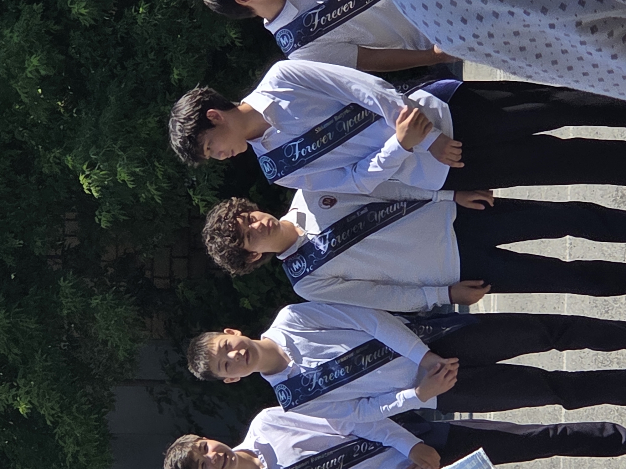 In an outdoor urban setting, possibly a schoolyard in Karaganda, Kazakhstan, five young males stand in a line, facing forward and slightly to the right. They appear to be students, likely dressed for an event or graduation, as each is wearing a white collared shirt and dark trousers. Over their shirts, they wear sashes with the inscription "Forever Young" and a stylized "M" emblem. The individuals display varying hairstyles and facial features. The background consists of lush green foliage, suggesting a well-maintained outdoor area. Sunlight illuminates the scene, casting subtle shadows. The ground beneath them appears to be a paved surface, possibly concrete or asphalt.