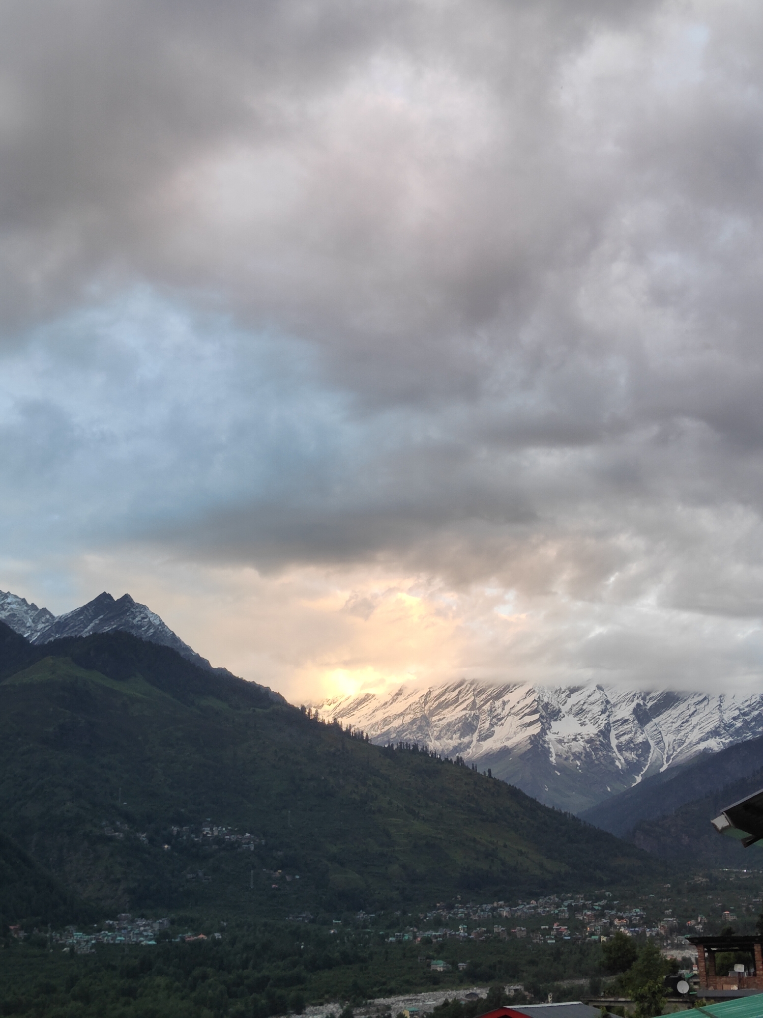 This image captures a panoramic view of the mountains and the town of Vashisht, India, under a dramatic, cloudy sky. The foreground features a dense forest covering the lower slopes of the mountains. Several small settlements, characterized by clusters of buildings with colorful rooftops, are visible nestled within the green hills. Further back, the majestic snow-capped peaks of the Himalayas rise sharply, their rugged terrain clearly defined against the sky. The sky itself is filled with heavy, grey clouds, but a patch of warm, golden light breaks through, illuminating a section of the snowy mountain range, suggesting either sunrise or sunset. The overall scene conveys a sense of natural grandeur and the peaceful coexistence of human settlements with the imposing mountain landscape.