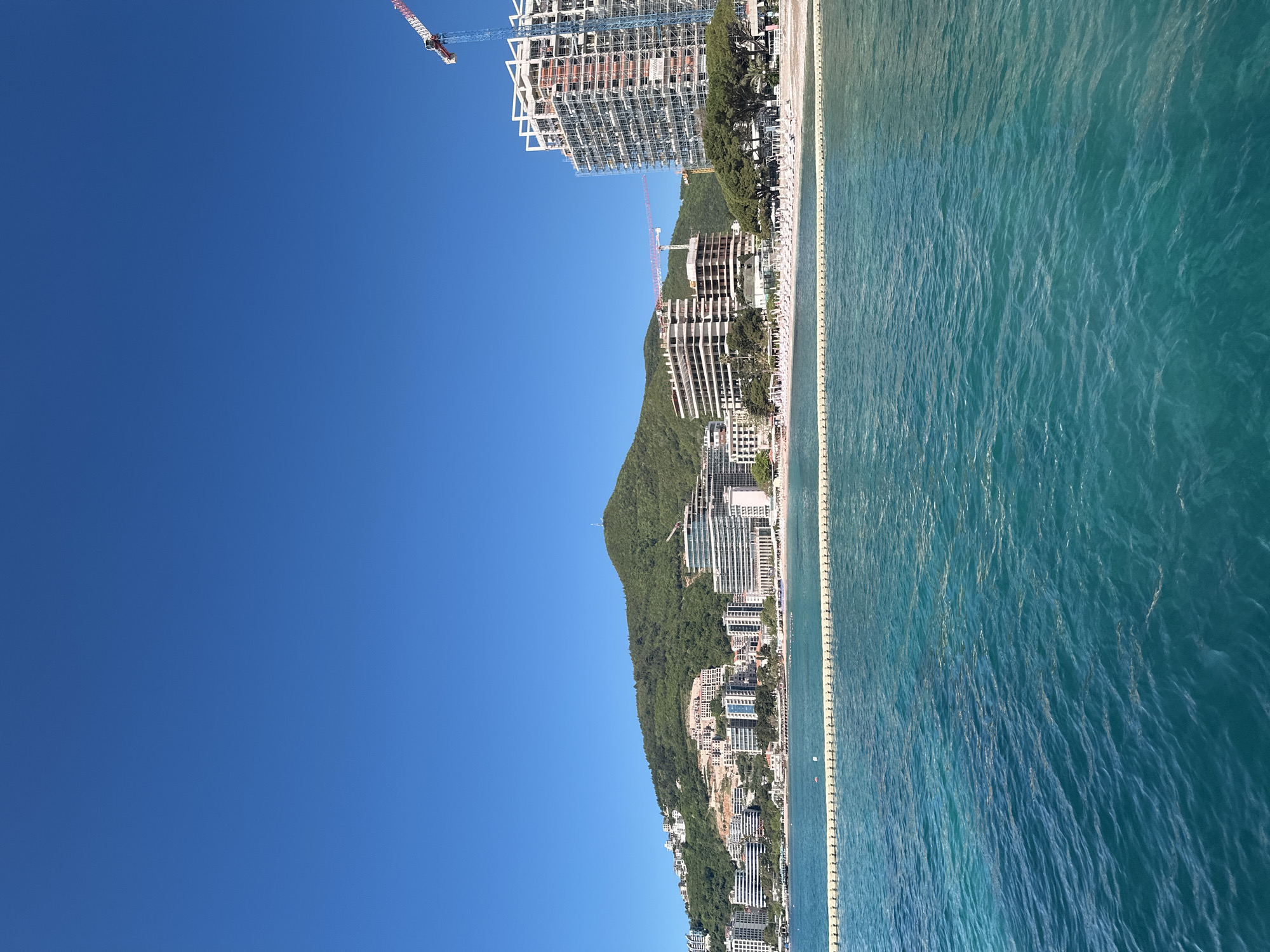 The image captures a coastal scene in Budva, Montenegro. The right side of the frame features the turquoise waters of the Adriatic Sea. Parallel to the sea is a protective barrier line.
The coastline is developed with high-rise buildings and residential complexes nestled against a green, mountainous backdrop. At the edge of the coastline are new buildings with construction cranes towering over them. The sky is clear blue.