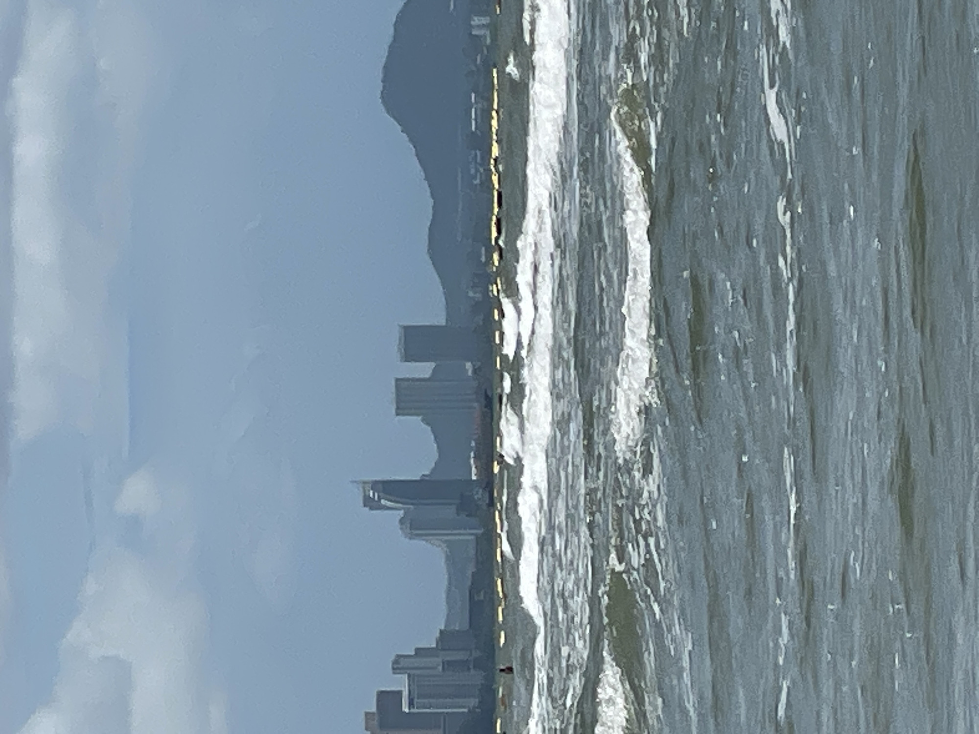 The image captures a wide-angle view of the sea in Pattaya City, Thailand, with a distant cityscape on the horizon. The foreground is dominated by choppy, grey-green ocean waves, some of which are breaking with white foam. In the middle ground, a line of yellow buoys stretches horizontally across the water, marking a boundary. A lone dark figure, possibly a person, is visible near the buoys on the left side of the frame. The background reveals a hazy skyline of tall buildings and mountains, characteristic of Pattaya's urban development along the coast. The sky above is overcast with muted tones of blue and white clouds.