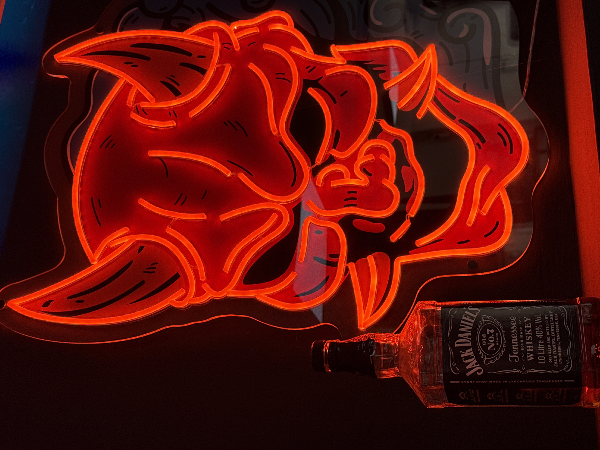 A bottle of Jack Daniel's Old No. 7 Tennessee Whiskey stands to the left of a large, red neon sign shaped like a demon's face. The sign is illuminated, casting a red glow on the bottle and the dark background. The demon face has horns, sharp teeth, and stylized eyes. The bottle is clear glass with a black label that features the Jack Daniel's logo and details about the whiskey. The scene is likely indoors, possibly in a bar or a personal collection space, given the neon sign. There is no indication of the city of Campillo Del Moro, Spain in the image.