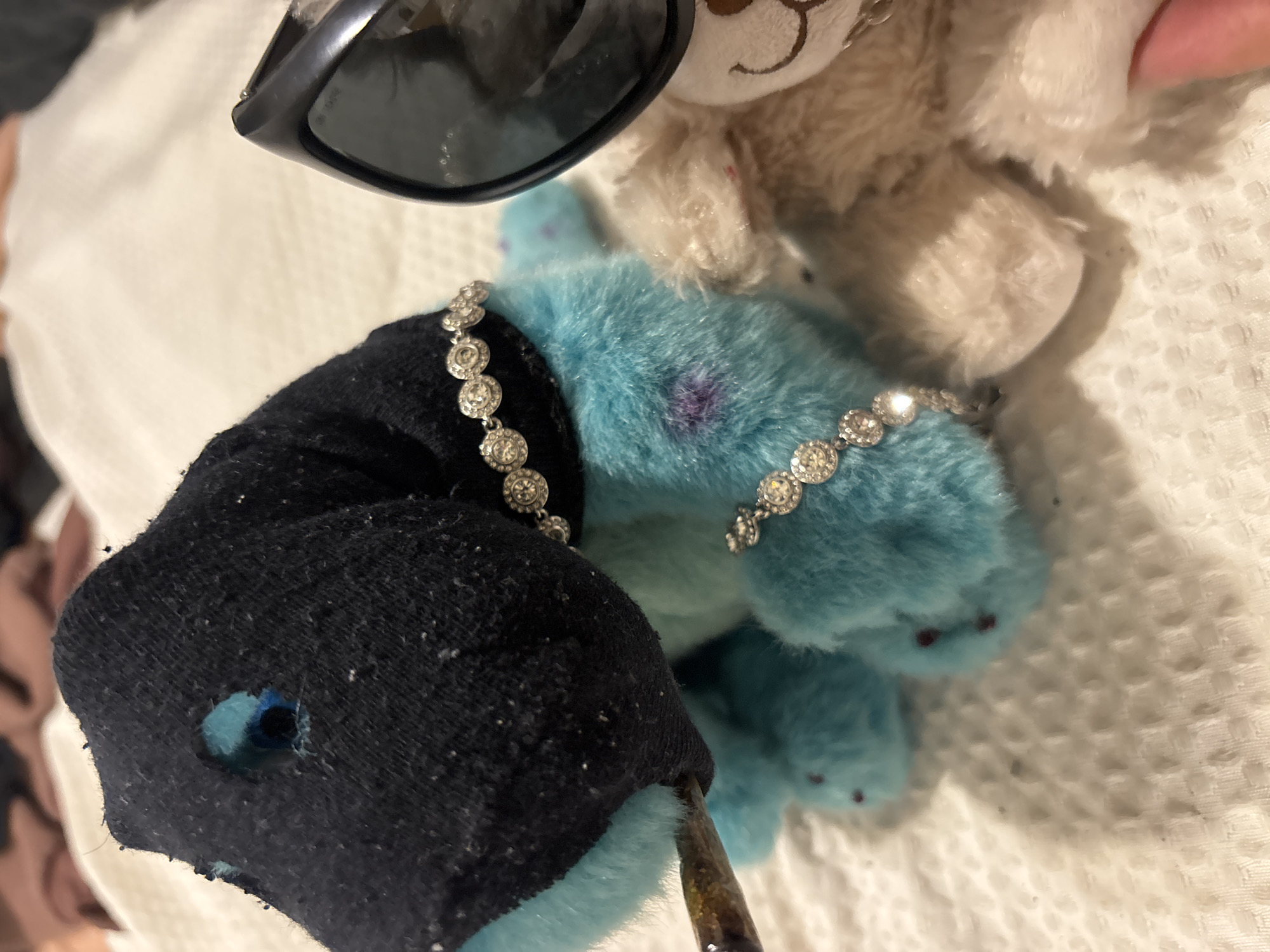The image presents a close-up, still-life scene with an assemblage of objects on what appears to be a white, textured fabric surface.
Foreground elements include a blue stuffed animal wearing a rhinestone bracelet, a black sock-like material with a hole revealing blue fur underneath, and what appears to be an oily tool or object touching the fur. The sock-like material also wears a rhinestone bracelet.
Background elements include a beige stuffed animal partially visible with sunglasses perched atop its head.
The image does not offer any information to pinpoint a location in Buenos Aires, Argentina, or any specific actions or interactions within a broader environment.