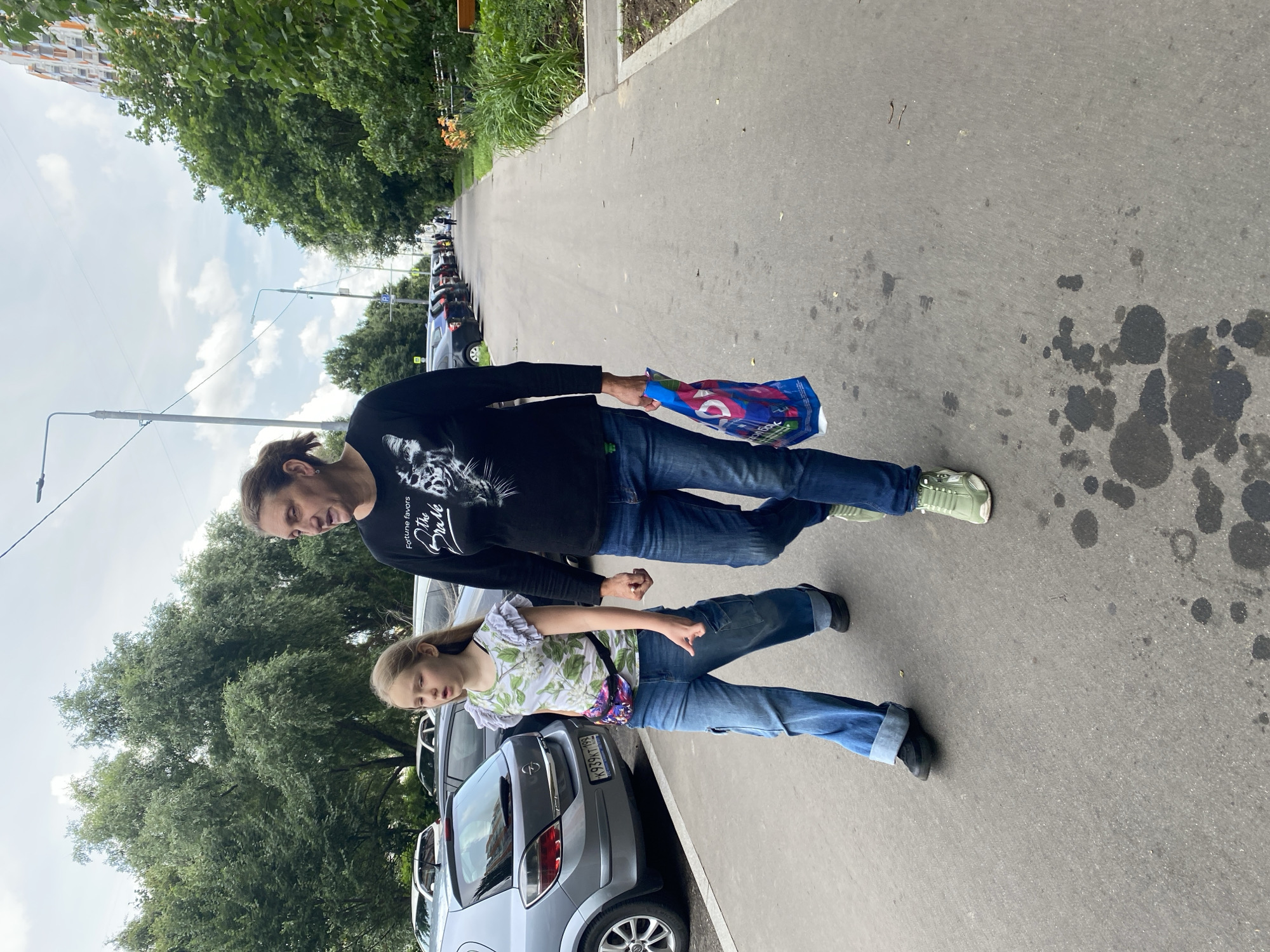 Two people, a woman and a young girl, are walking on a sidewalk in Khimki, Russia. The woman is on the right, wearing a black long-sleeved shirt with a graphic print and blue jeans. She is holding a blue shopping bag in her right hand. The girl is on the left, wearing a white floral print t-shirt with ruffled sleeves and light blue wide-leg jeans. She has a fanny pack worn across her chest. They are walking on a paved sidewalk next to a parking lot filled with cars on the left and a strip of grass and trees on the right. The sky is cloudy.