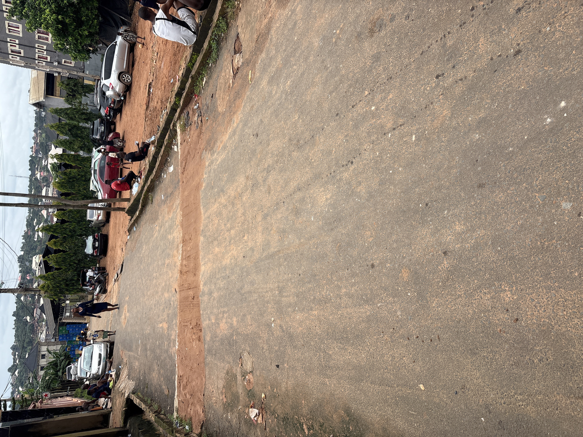 This is a medium shot, taken from a slightly elevated position, of a street scene in Nkpor, Nigeria. The street is paved with asphalt and is noticeably dirty, with patches of red earth and debris scattered across its surface. On the left side of the street, a building with an exposed brick facade is visible, with people sitting on a ledge in front of it. Further down the street, several cars are parked on a dirt lot to the right, including a silver SUV, a red sedan, and a black car. A group of people are gathered around the cars, with some sitting on the ground and others standing. A young boy is playing with a soccer ball in the dirt lot. In the background, more buildings and trees are visible, suggesting a densely populated urban area. The sky is overcast. A woman in a dark dress is walking away from the viewer up the street, while a young boy in a white and yellow jersey walks towards her. A motorcycle is parked near some of the cars.