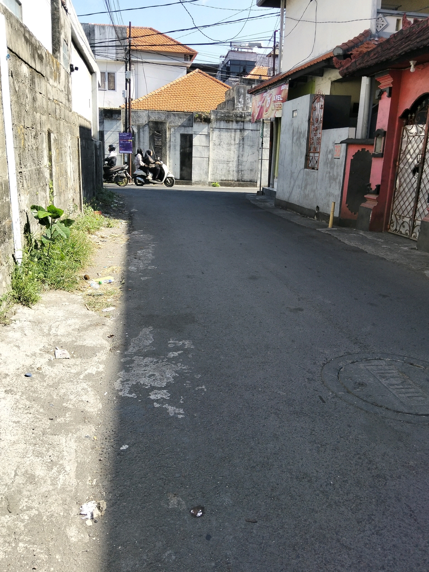 The image depicts a street scene in Kuta, Indonesia, characterized by buildings with traditional orange tile roofs and concrete walls. In the foreground, a paved road is visible, with a portion of the sidewalk on the left side showing patches of dry grass and scattered debris, including what appear to be paper wrappers and plastic bottles. A young green plant grows near the wall on the left. In the middle ground, two motorbikes are parked. On the first motorbike, one person is seated and a second person is standing next to it. On the second motorbike, two individuals are riding, one driving and the other as a passenger. The individuals on both motorbikes are wearing helmets. Buildings line both sides of the street, with one building on the right featuring a pink facade and a decorative metal gate. Overhead, a tangle of electrical wires is suspended between poles. The sky is clear and blue.