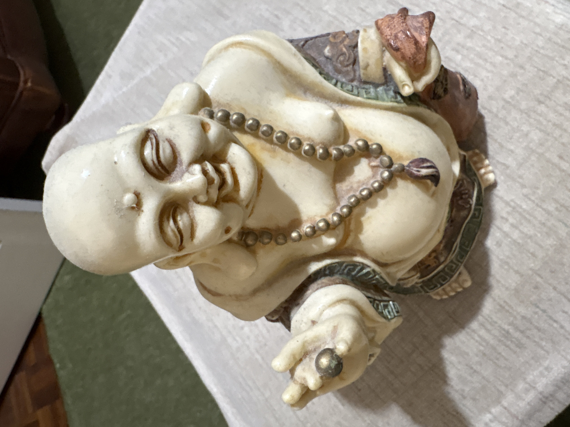 A close-up, high-angle shot shows a cream-colored statue of a laughing Buddha. The statue is adorned with a beaded necklace and sits on a textured, light-colored surface. The background is blurred, showing a hint of a wooden floor and a green rug. The statue's features are detailed, with closed eyes, a smiling mouth, and a round belly. Its right hand is extended, holding a small, round object. The statue's clothing is depicted with patterns and textured fabric. The lighting is soft, casting a subtle shadow to the left of the statue. This image does not contain enough information to determine its location in the city of Chelmsford, United Kingdom, or any other specific geographic details.