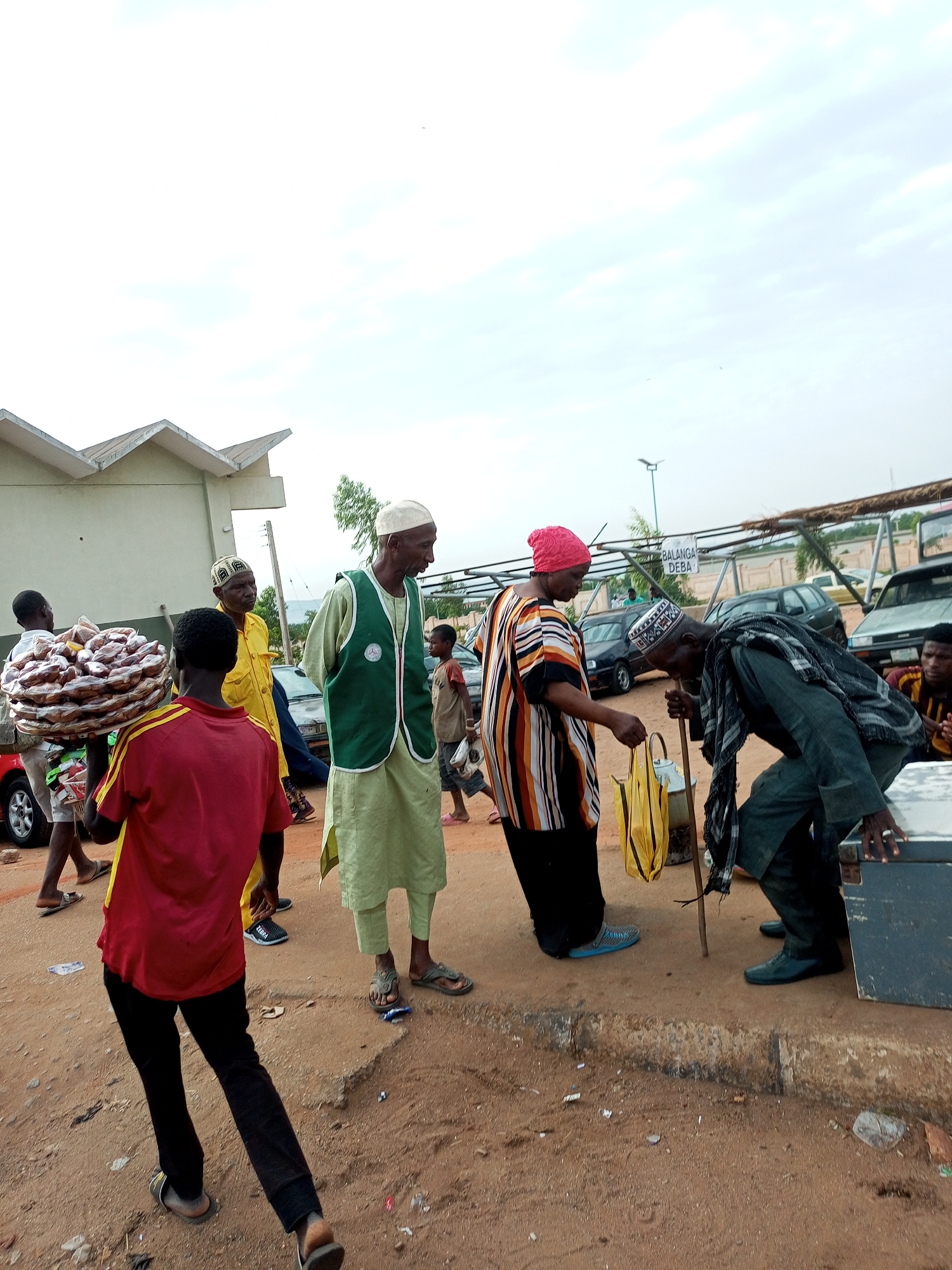 A group of people are gathered on a dusty, unpaved street in Gombe, Nigeria. In the foreground, a young person in a red shirt and black pants carries a large tray of food items on their head. To their right, an older man wearing a green vest over a light green traditional outfit stands with his hands behind his back. Further to the right, an older man wearing a green scarf and dark pants is bending over and appears to be interacting with a woman wearing a brightly colored striped shirt and a pink headscarf. The woman is holding a yellow bag and a cane. In the background, several vehicles, including cars and a truck, are parked. A white building with a distinctive angular roofline is visible on the left side of the image. A sign with text that appears to be "BALANGA DEBA" is mounted on poles in the background. The sky is overcast.