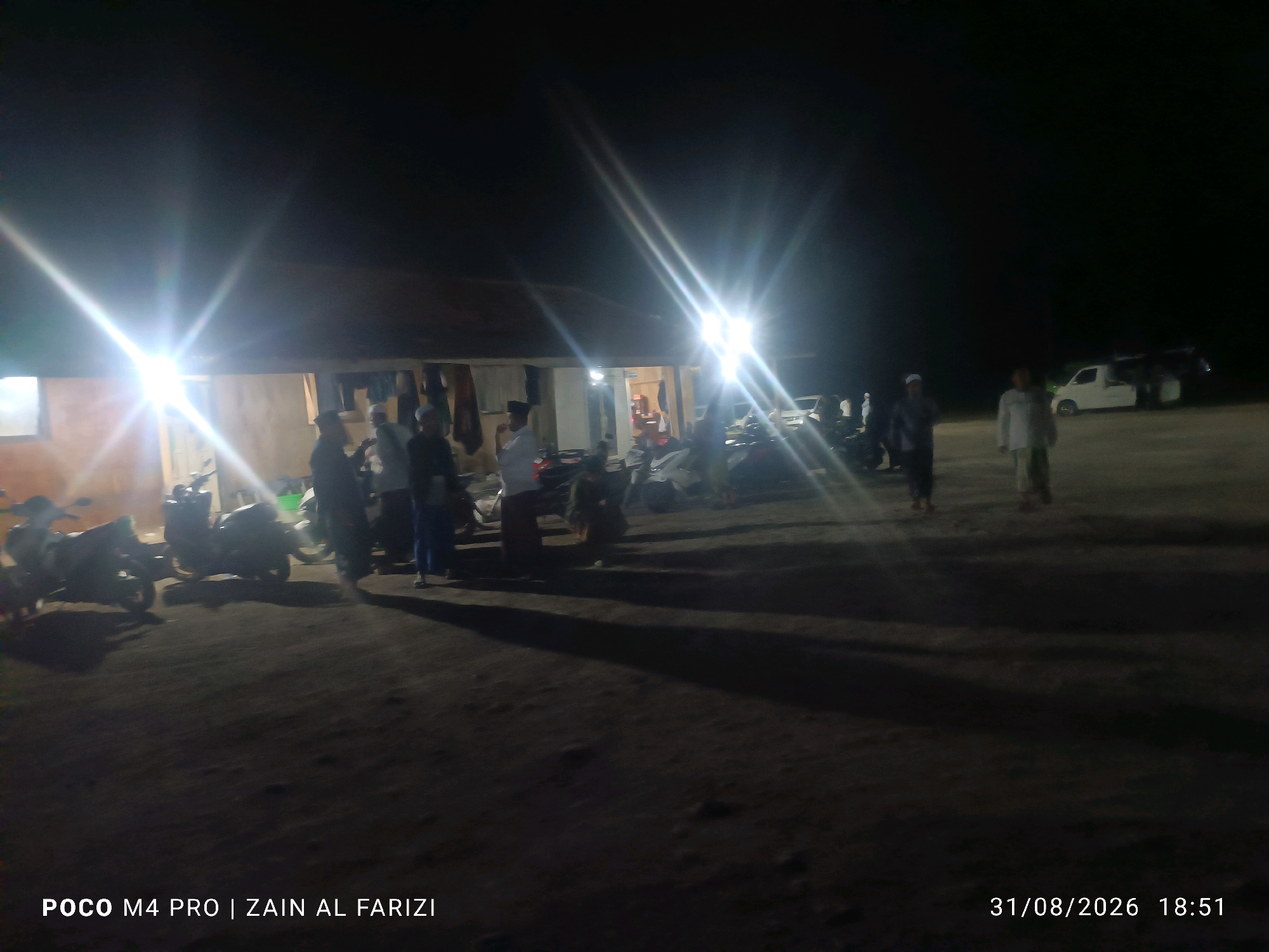 The image captures an outdoor scene at night, likely in Bangkalan, Indonesia, as indicated by the timestamp and the POCO M4 PRO watermark which is a phone model. In the foreground, the ground is dark and appears to be unpaved. Several motorcycles are parked, some casting shadows. A low-lying building with a dark roof is visible in the mid-ground, with lights emanating from it, suggesting it's open or illuminated. Some clothing items are hanging outside the building. A few individuals are standing around the building and near the motorcycles. Further in the background, a white van and another vehicle are parked. The lighting is artificial, with strong glare from light sources creating lens flares and bright streaks across the image, which is typical of night-time photography in areas with direct light sources. The overall impression is a gathering or activity taking place outside a building at night, with vehicles present.